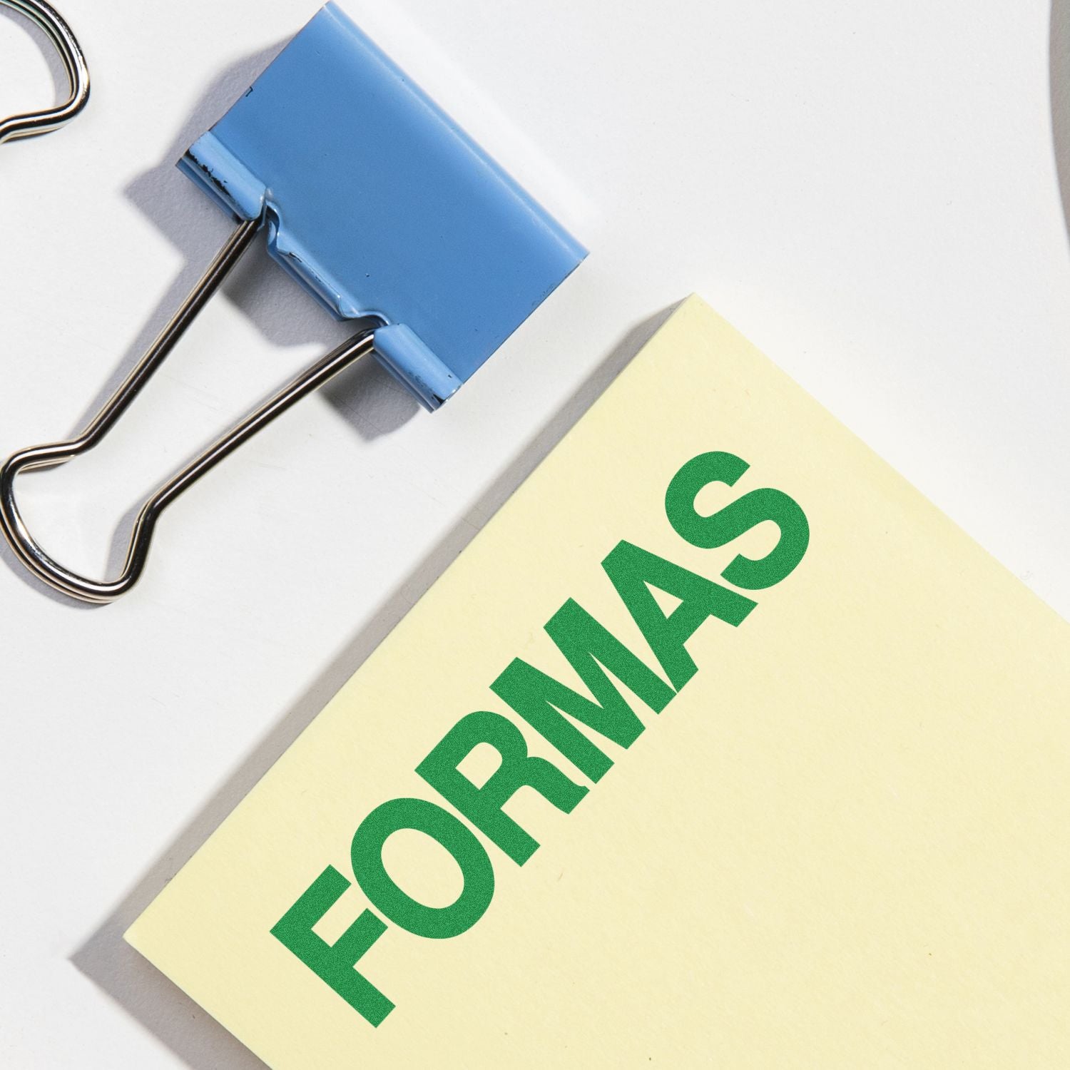 Large Self Inking Formas Stamp in use, stamping FORMAS in green on a yellow paper, next to a blue binder clip on a white surface.