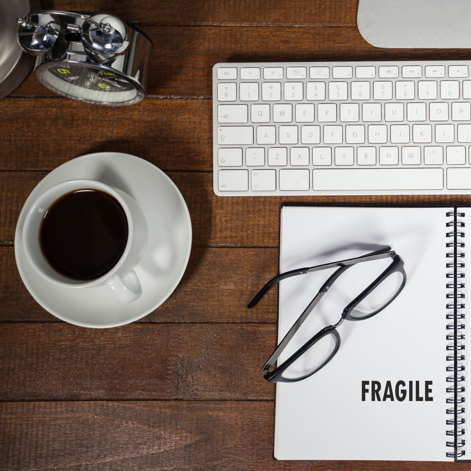 Slim Pre-Inked Fragile Stamp on a notebook beside glasses, a coffee cup, a keyboard, and a clock on a wooden desk.