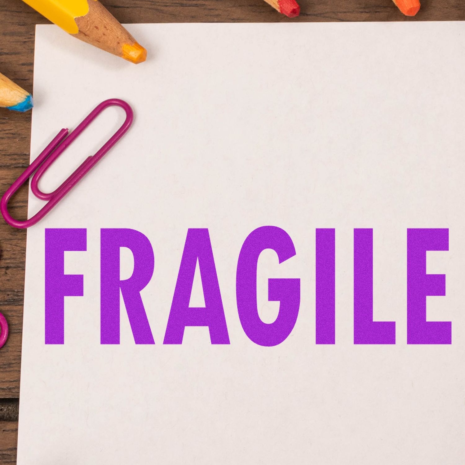 Large Self Inking Fragile Stamp in use, stamping FRAGILE in bold purple letters on a white paper, surrounded by colorful pencils and a paperclip.