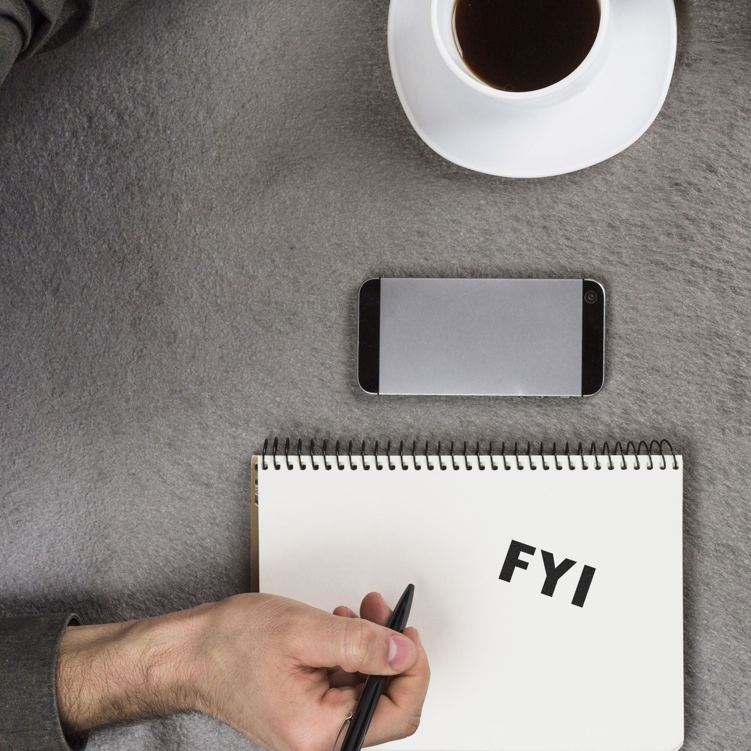 A hand holding a pen next to a notebook stamped with FYI using the FYI Rubber Stamp, with a smartphone and a cup of coffee nearby.