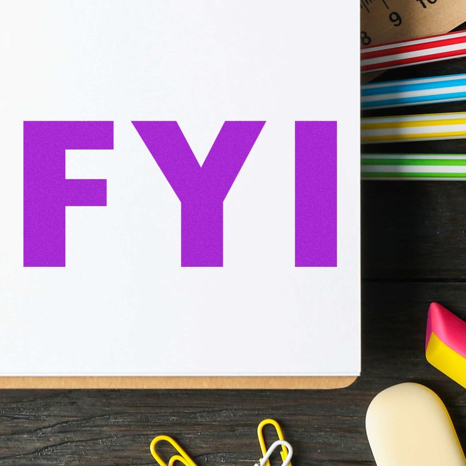 FYI Rubber Stamp in use on a white paper, surrounded by colorful paper clips, a ruler, and other office supplies on a wooden desk.