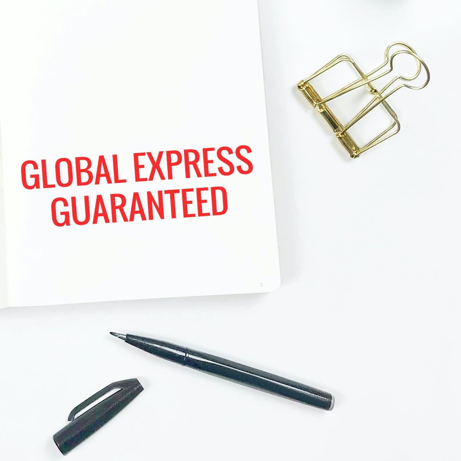 Self Inking Global Express Guaranteed Stamp in red ink on white paper, with a black pen and gold paper clip nearby on a white surface.