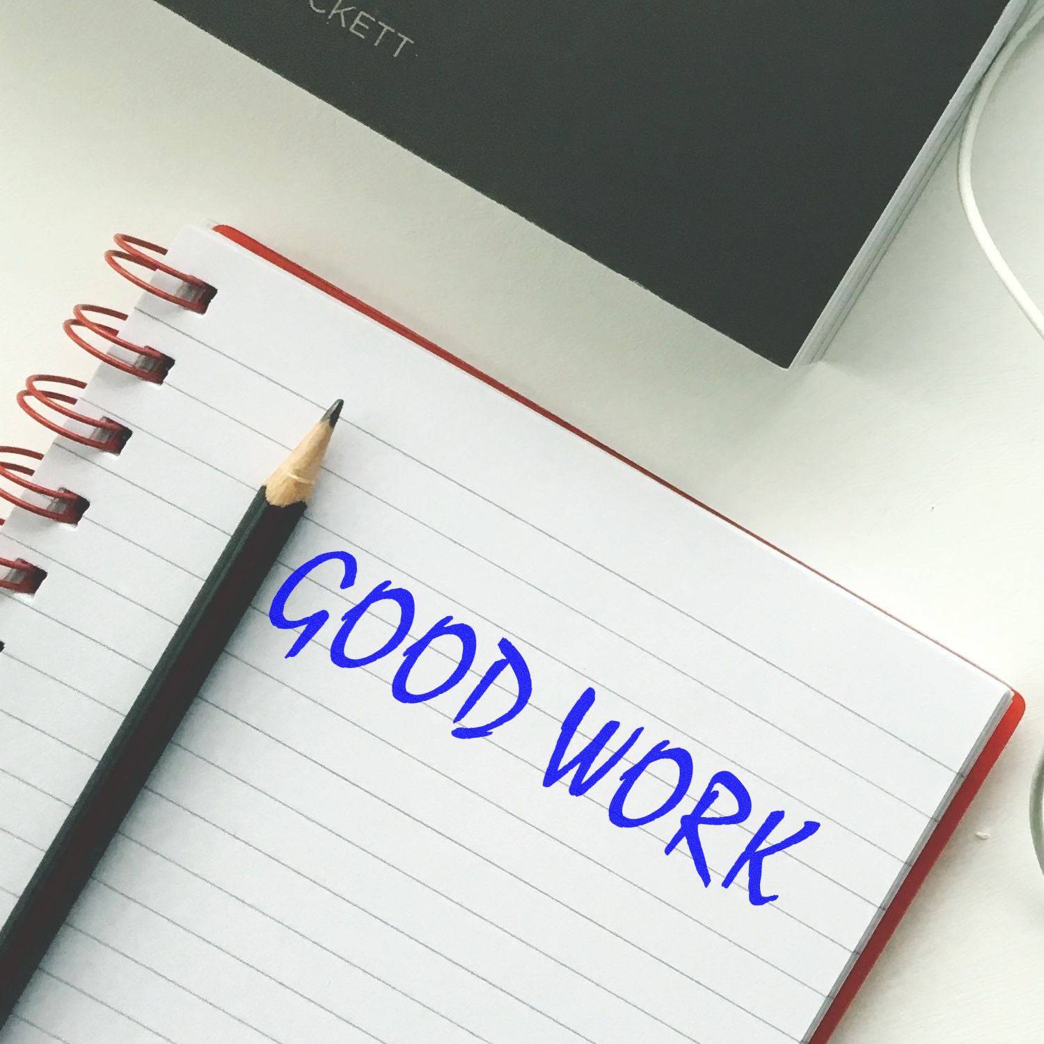 A notebook with GOOD WORK stamped in blue ink using the Large Pre-Inked Good Work Stamp, next to a pencil on a white desk.