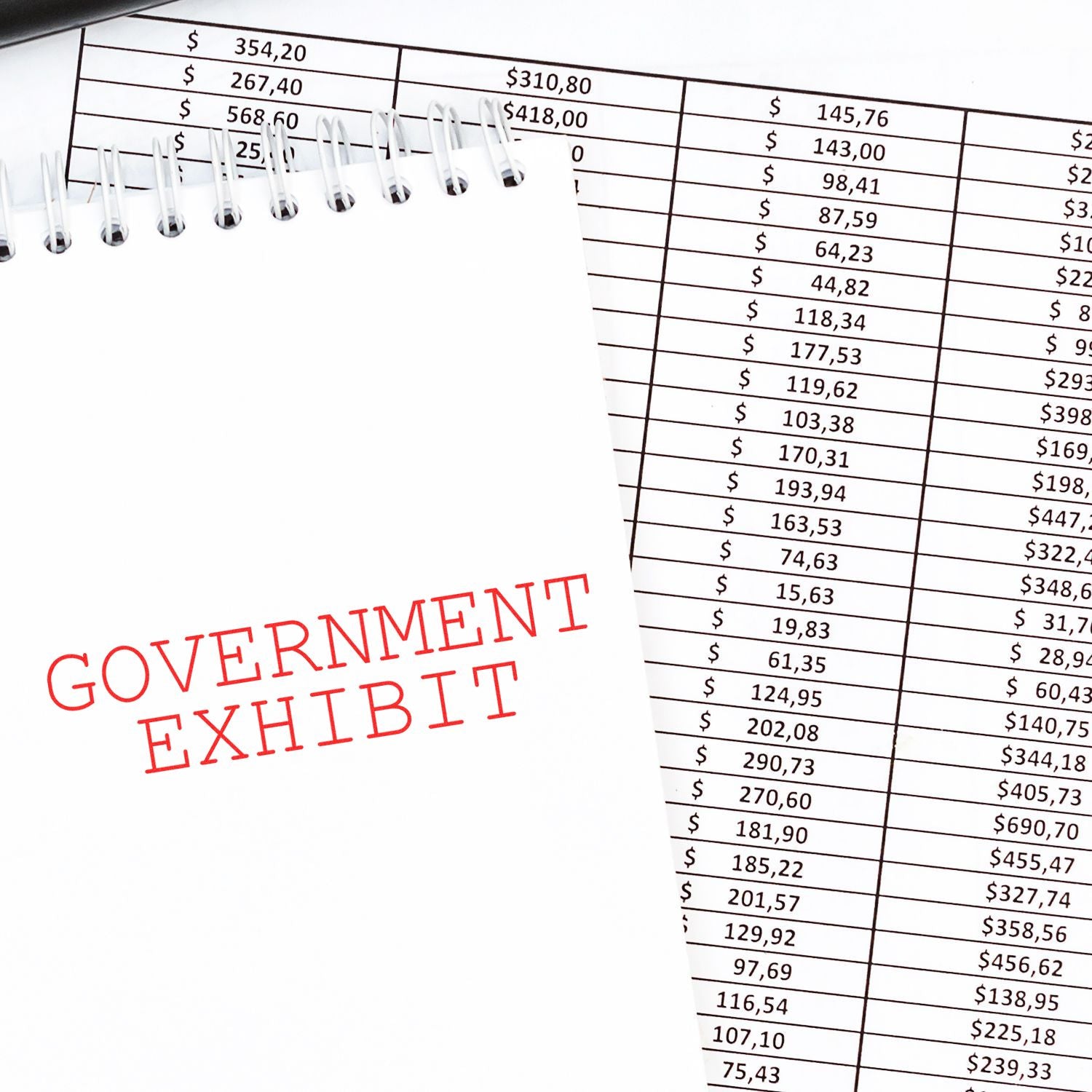 Slim Pre-Inked Government Exhibit Stamp used on a notebook page, placed over a financial spreadsheet.