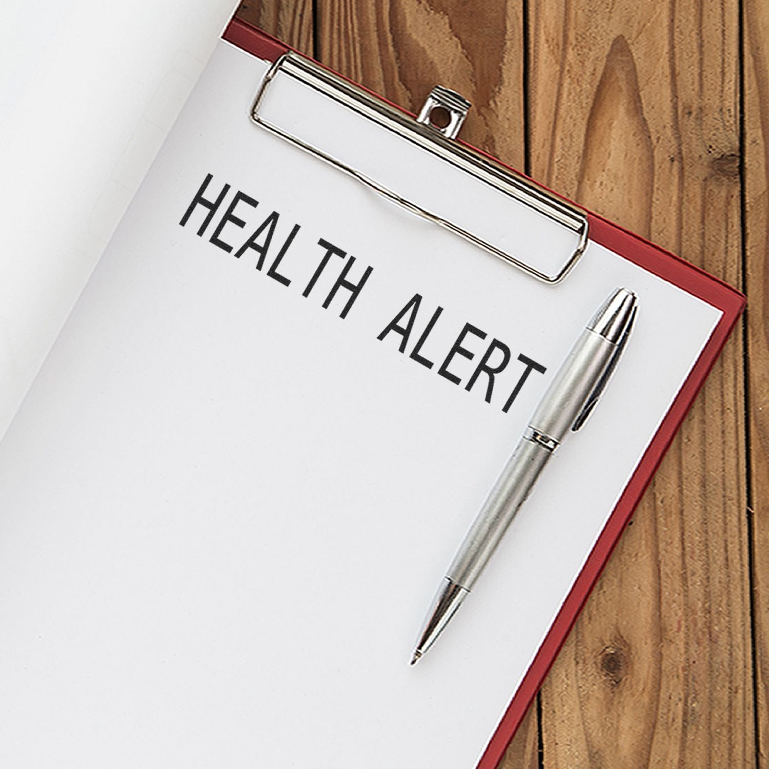 Clipboard with paper stamped HEALTH ALERT using the Health Alert Rubber Stamp, with a silver pen placed beside it on a wooden surface.