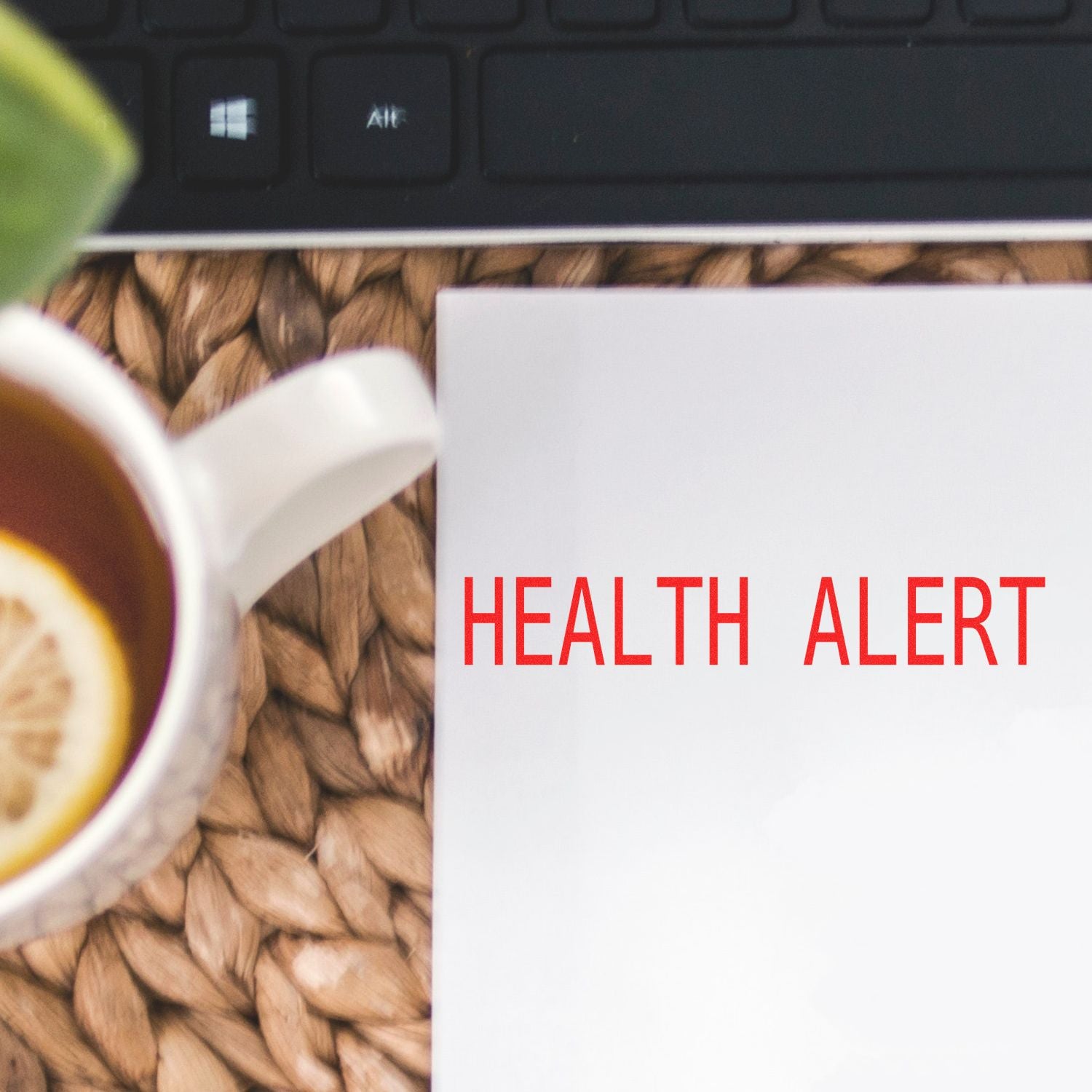 Health Alert Rubber Stamp in red ink on white paper, next to a cup of tea with lemon and a keyboard on a woven mat.