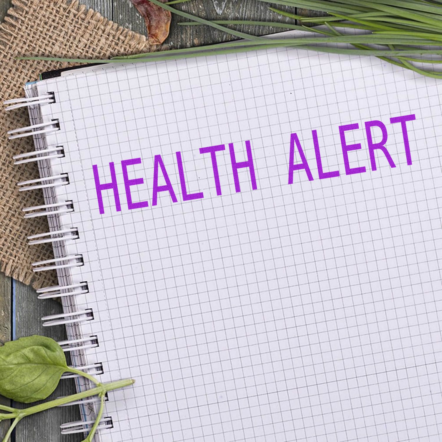 Slim Pre-Inked Health Alert Stamp used on a grid notebook, surrounded by green leaves and a burlap fabric on a wooden surface.