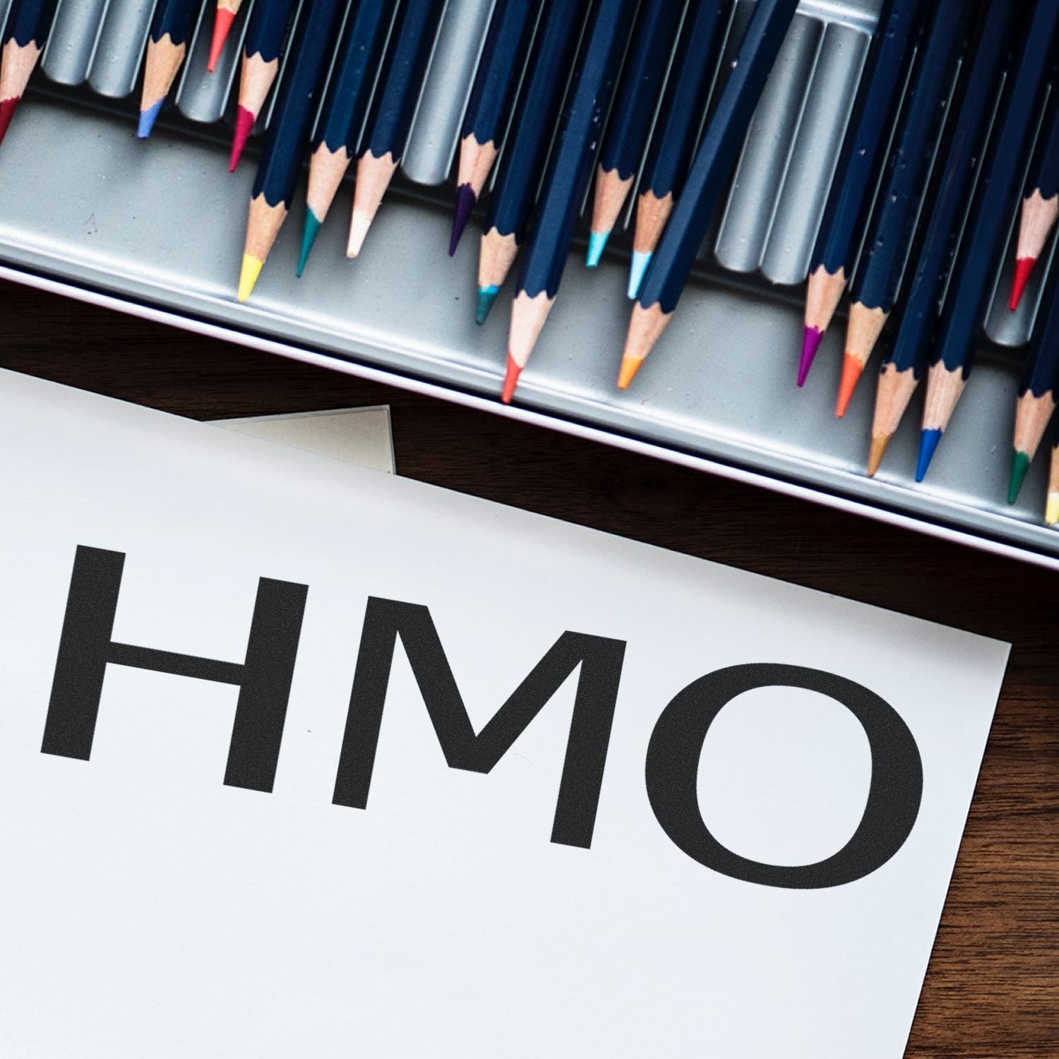 Self Inking HMO Stamp in use on white paper, surrounded by colorful pencils in a metal tray on a wooden surface.