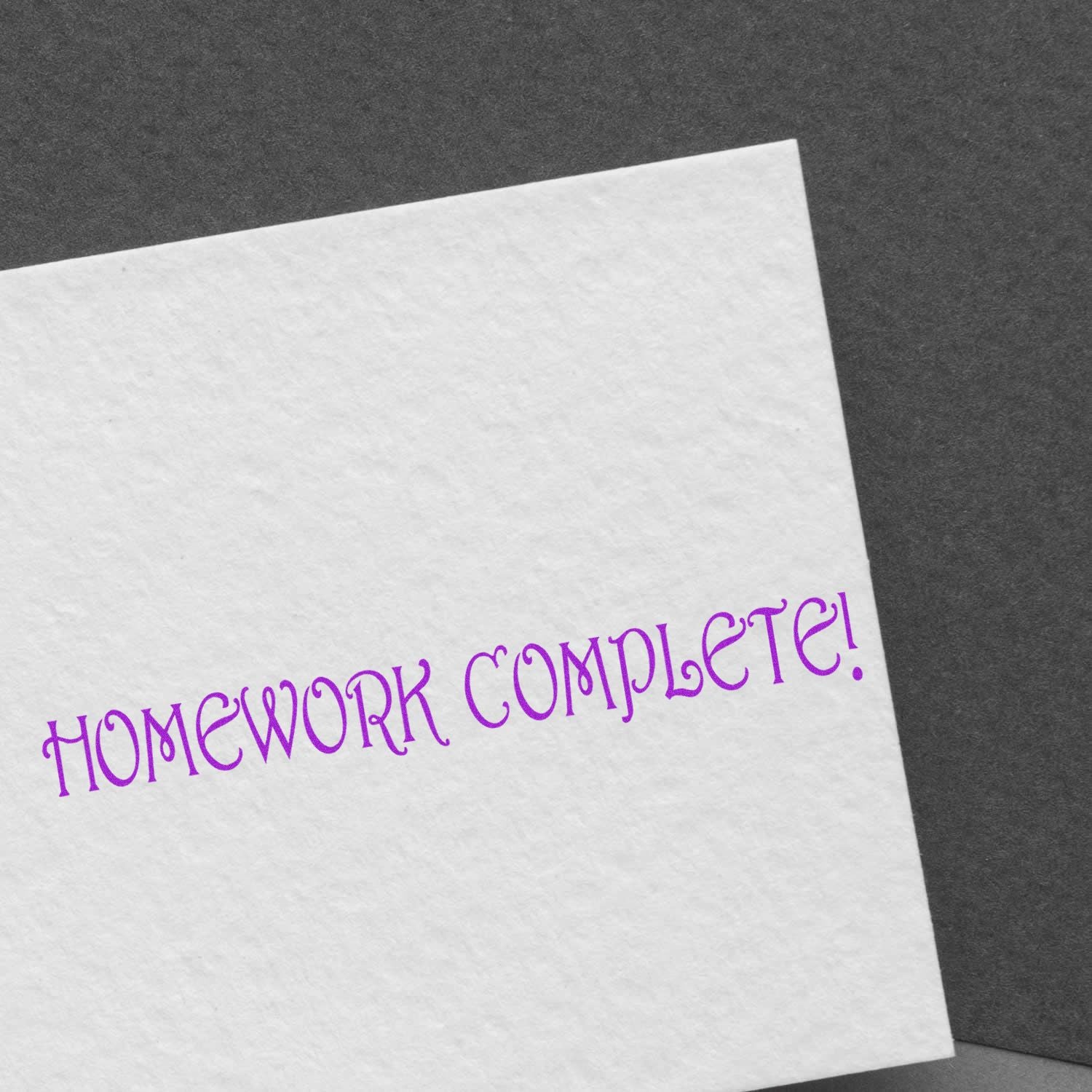 A white paper stamped with HOMEWORK COMPLETE! in purple ink using the Homework Complete Rubber Stamp, placed on a dark background.