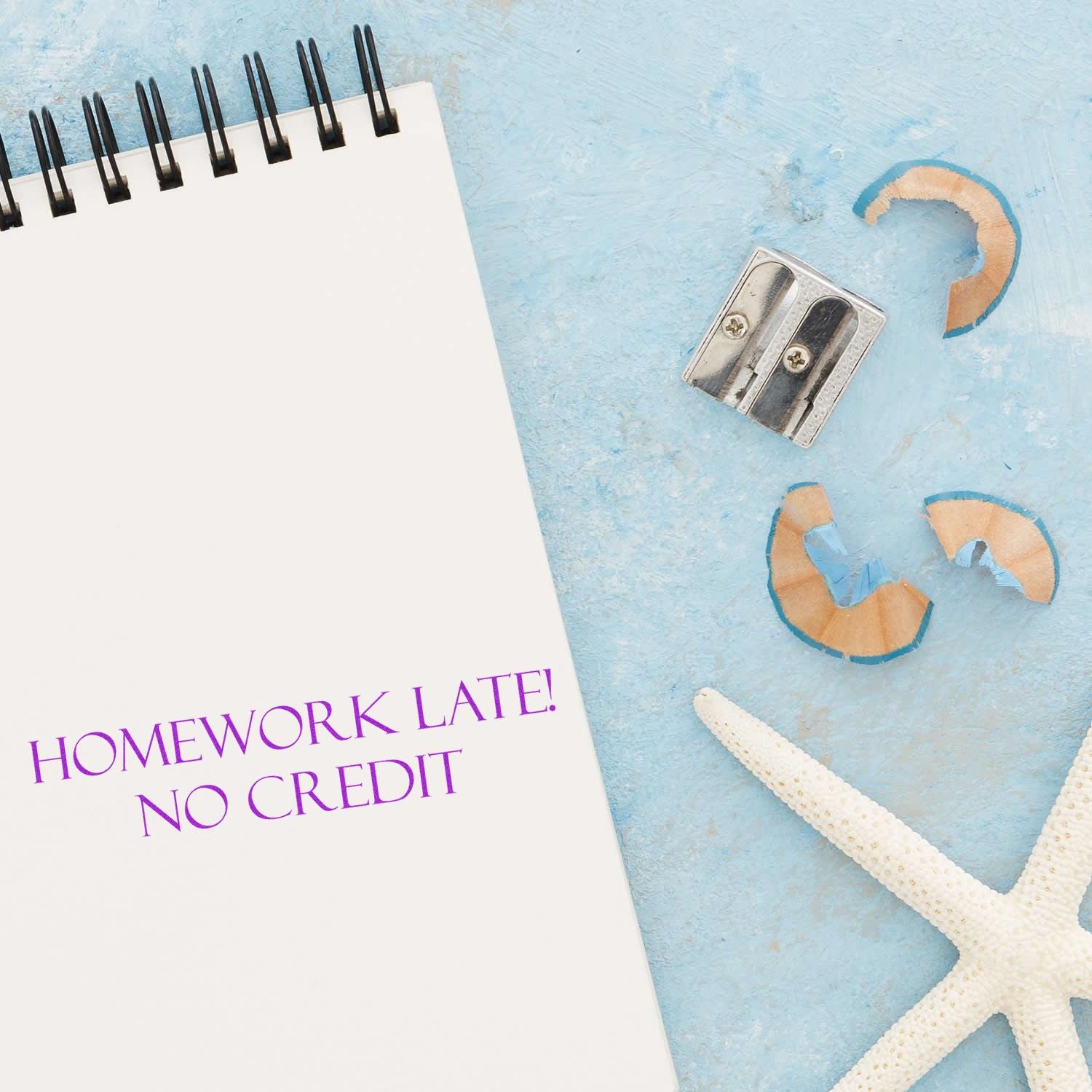 A Homework Late No Credit Rubber Stamp is used on a notebook page, surrounded by a pencil sharpener, shavings, and a starfish on a blue surface.