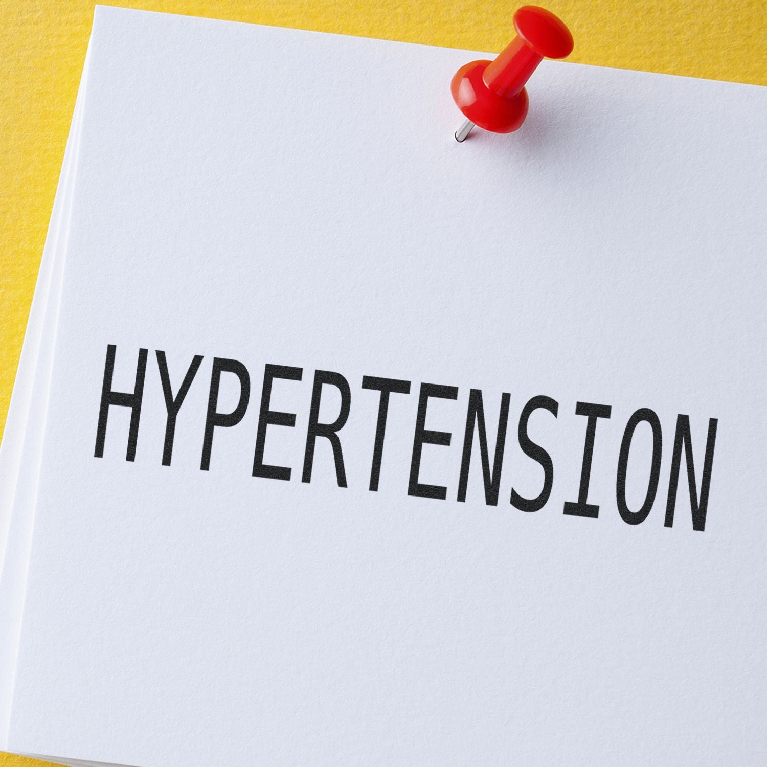 A red pushpin holds a white card stamped with HYPERTENSION using the Large Pre-Inked Hypertension Stamp against a yellow background.