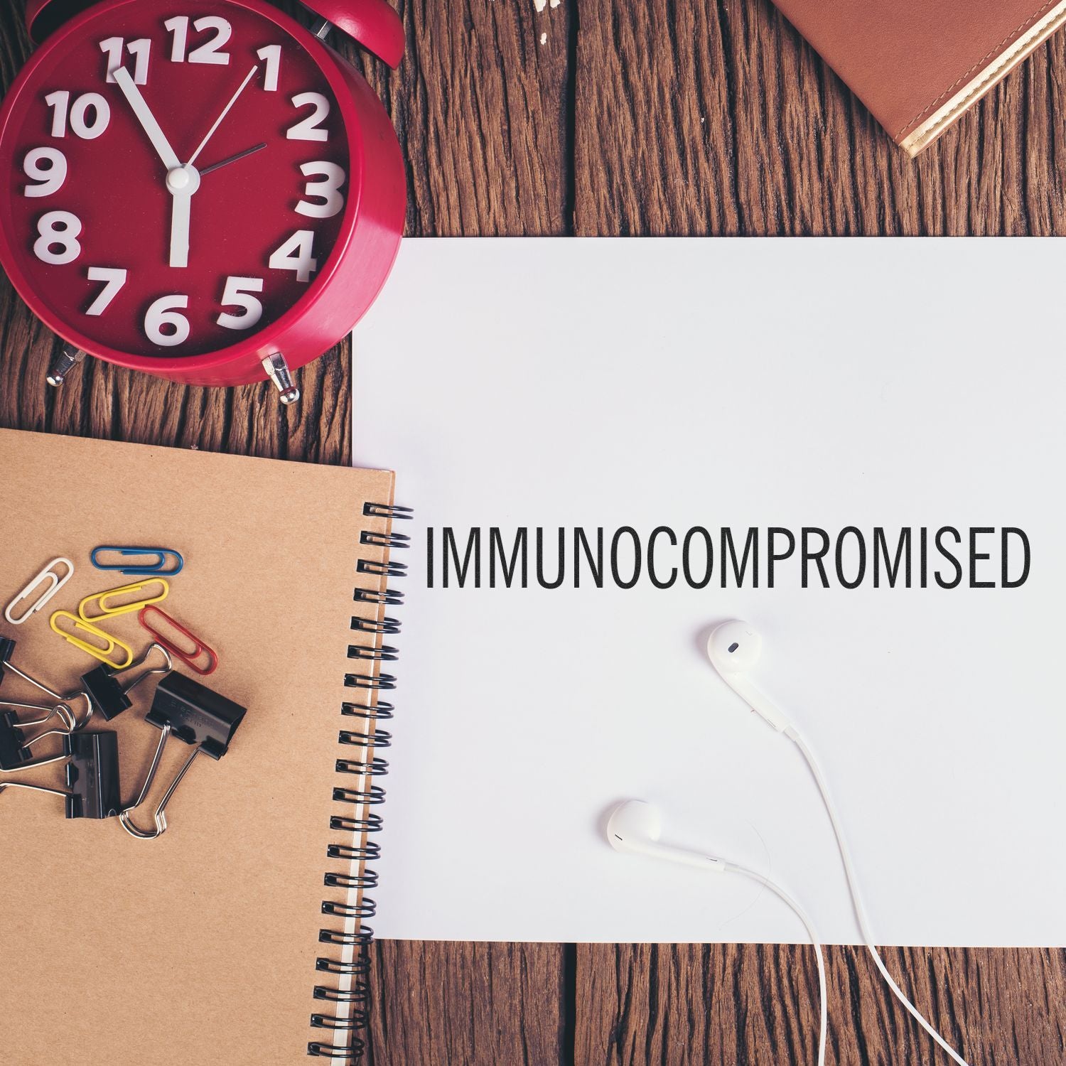 Large Pre-Inked Immunocompromised Stamp on white paper, surrounded by a red clock, notebook, paper clips, and earphones on a wooden desk.