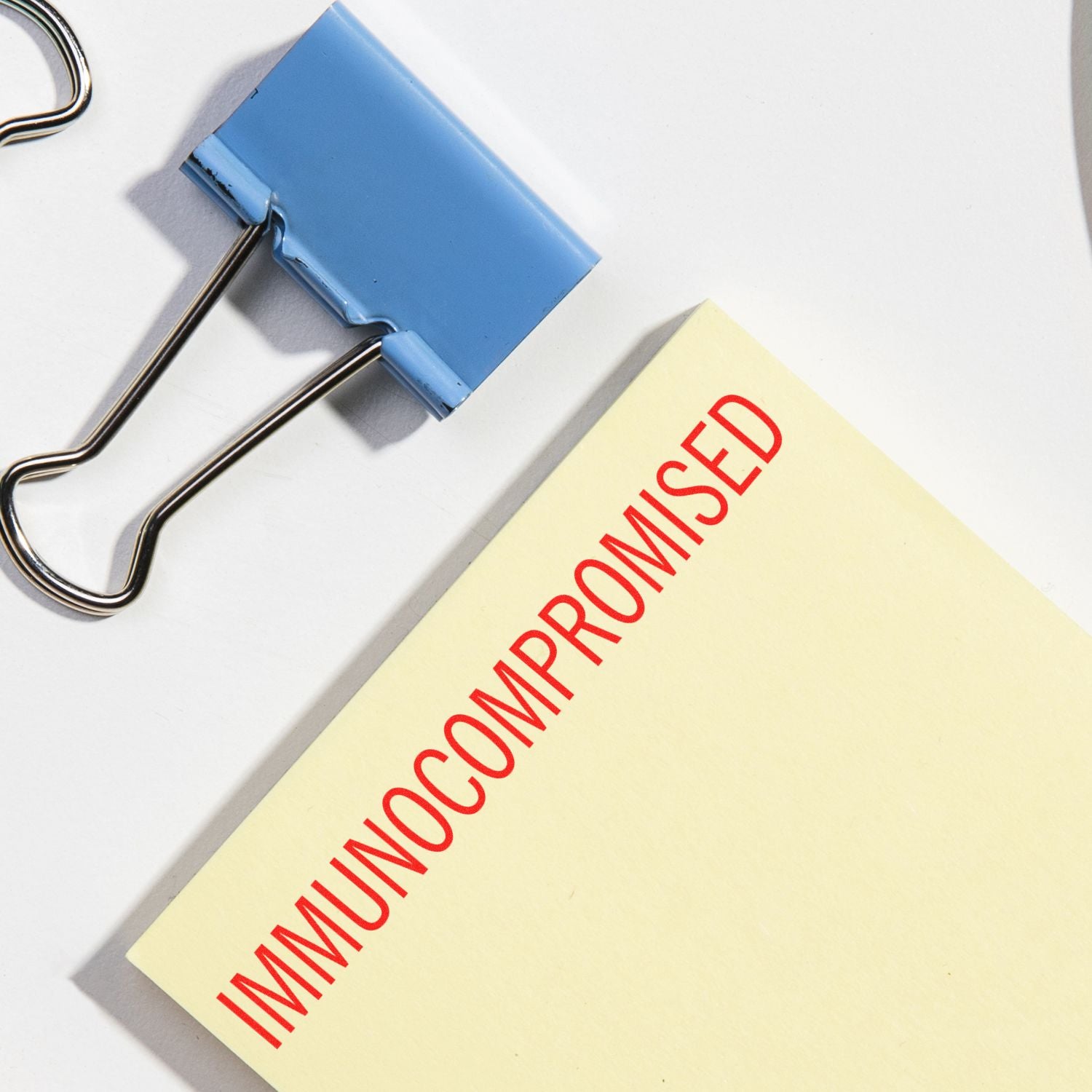 Large Pre-Inked Immunocompromised Stamp in red ink on a yellow paper, next to a blue binder clip on a white surface.