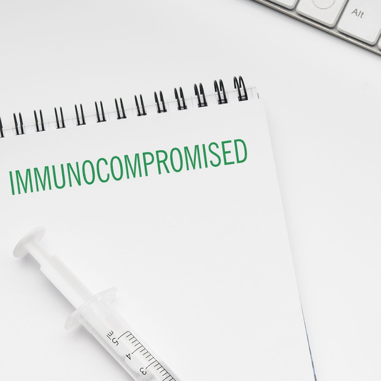 Slim Pre-Inked Immunocompromised Stamp marking a notebook with IMMUNOCOMPROMISED in green, next to a syringe and keyboard.