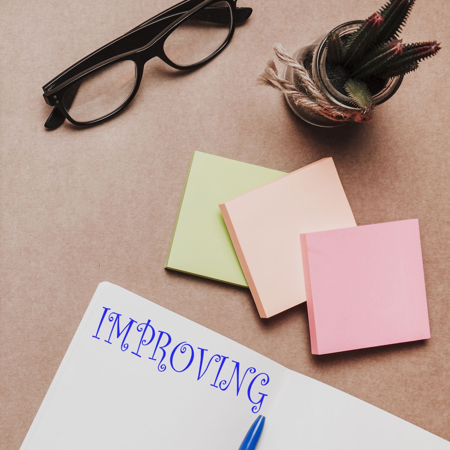 Notebook stamped with Improving using the Improving Rubber Stamp, surrounded by sticky notes, glasses, and a potted plant on a desk.