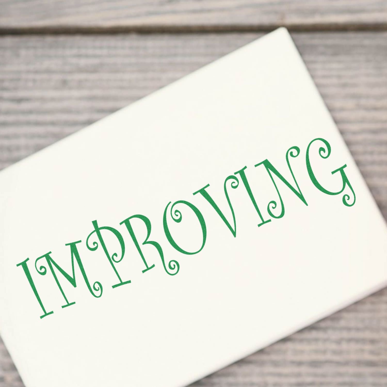 A white paper stamped with the word IMPROVING in green ink using the Self Inking Improving Stamp, placed on a wooden surface.