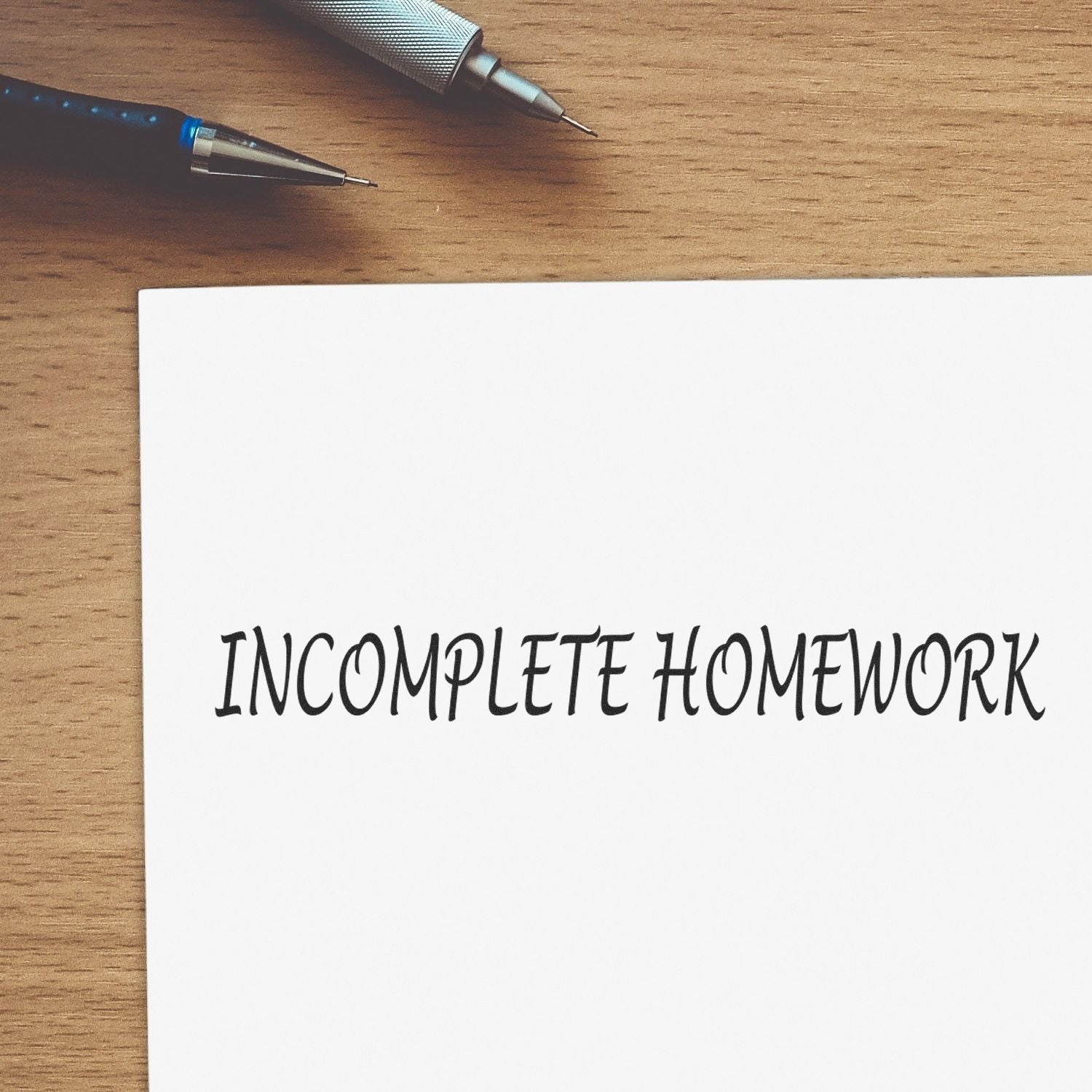 Large Incomplete Homework Rubber Stamp on a white paper with two pens on a wooden desk.