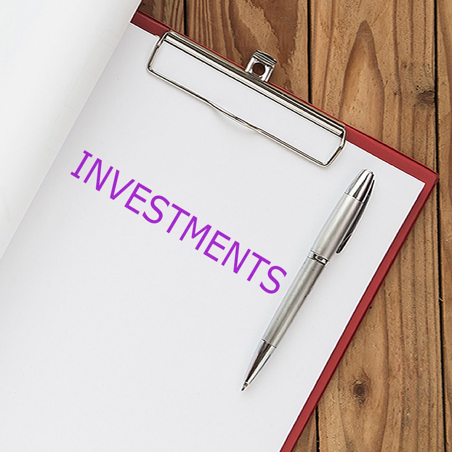 Clipboard with a paper stamped INVESTMENTS in purple ink, using the Large Investments Rubber Stamp, next to a silver pen on a wooden surface.