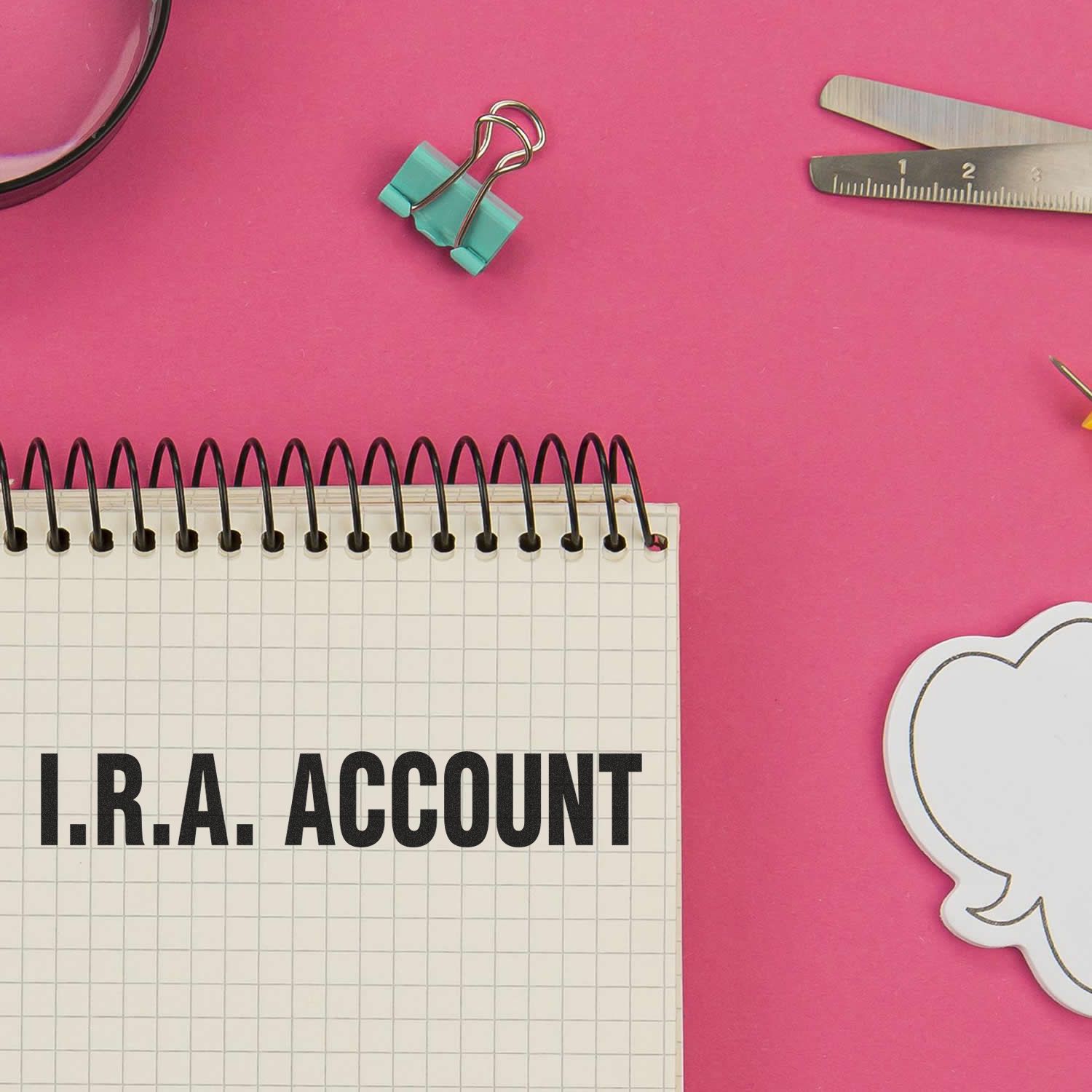 A spiral notebook with I.R.A. ACCOUNT stamped on it, surrounded by office supplies on a pink background.