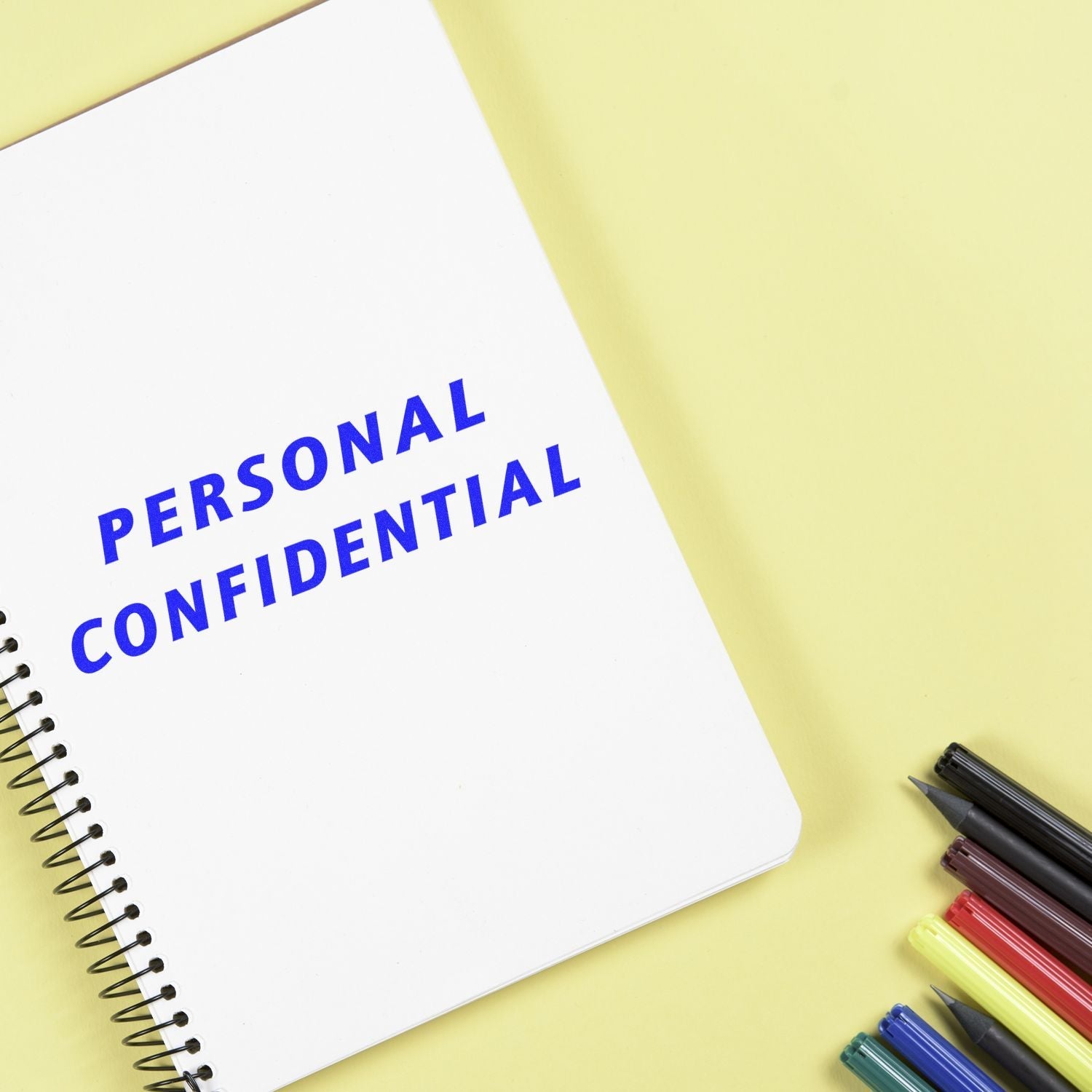 A notebook stamped with PERSONAL CONFIDENTIAL using the Self Inking Italic Personal Confidential Stamp, surrounded by colorful pens on a yellow background.