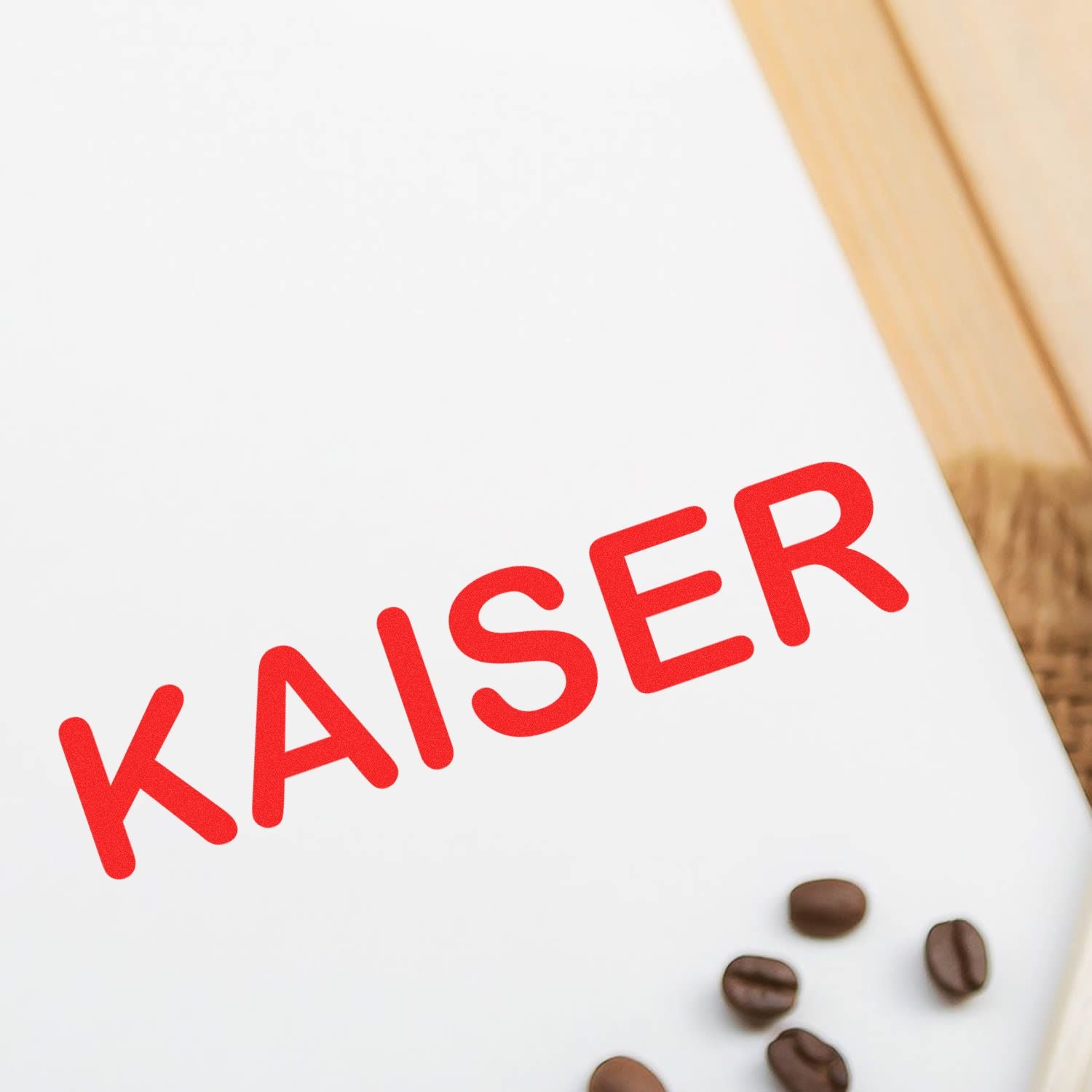 Large Self Inking Kaiser Stamp in use, imprinting the word KAISER in red ink on a white surface, with coffee beans scattered nearby.