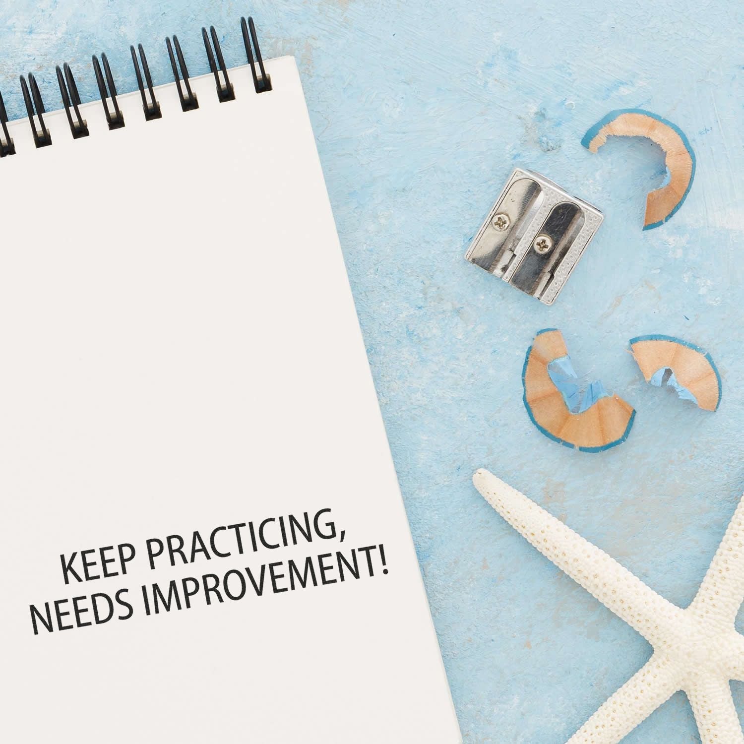 A Keep Practicing Needs Improvement Rubber Stamp is shown next to a notebook, pencil sharpener, pencil shavings, and a starfish on a blue surface.