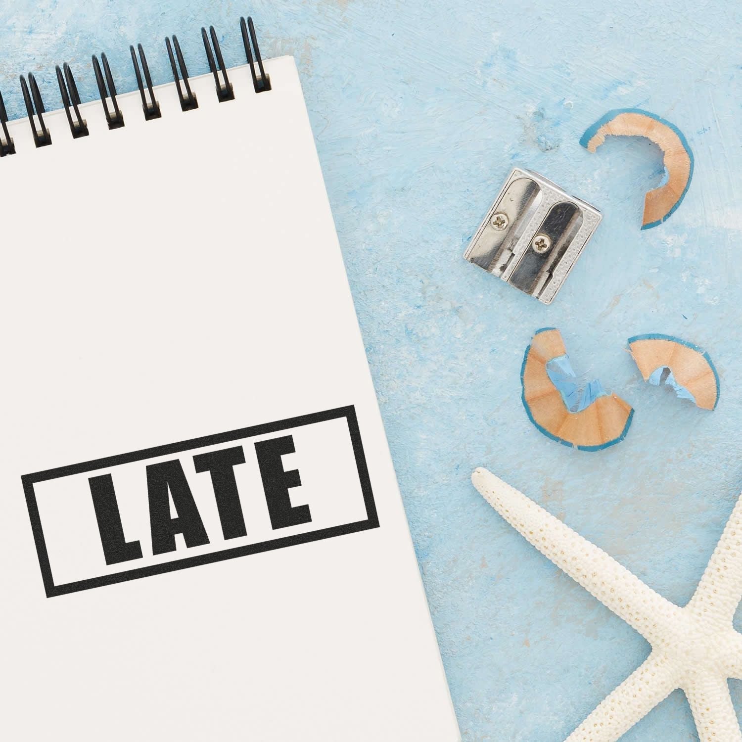 Notebook stamped with LATE using Self Inking Late with Border Stamp, placed on a blue surface with a sharpener, pencil shavings, and a starfish.