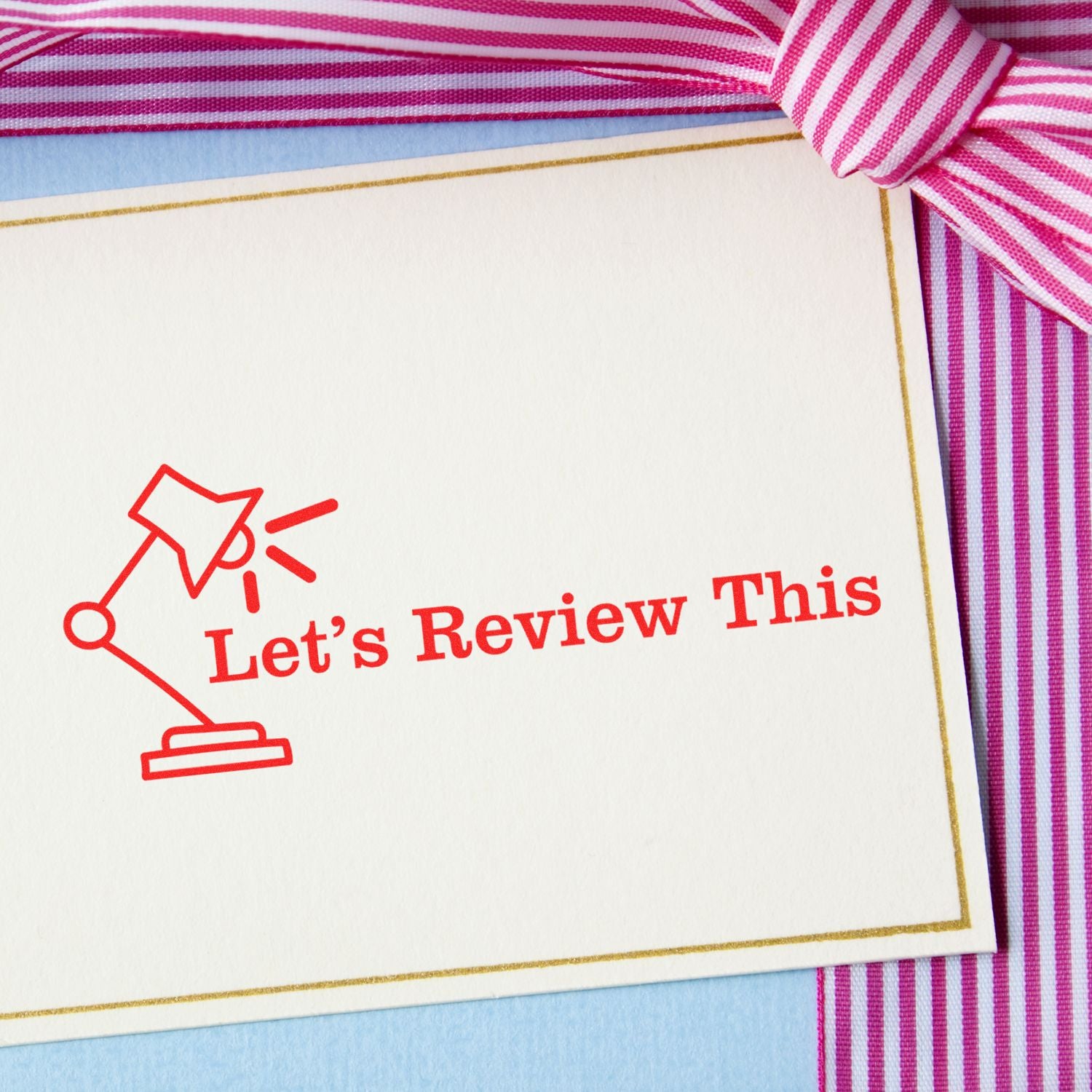 Let's Review This with Lamp Rubber Stamp on a white card with red text, placed on a striped pink and white background with a bow.