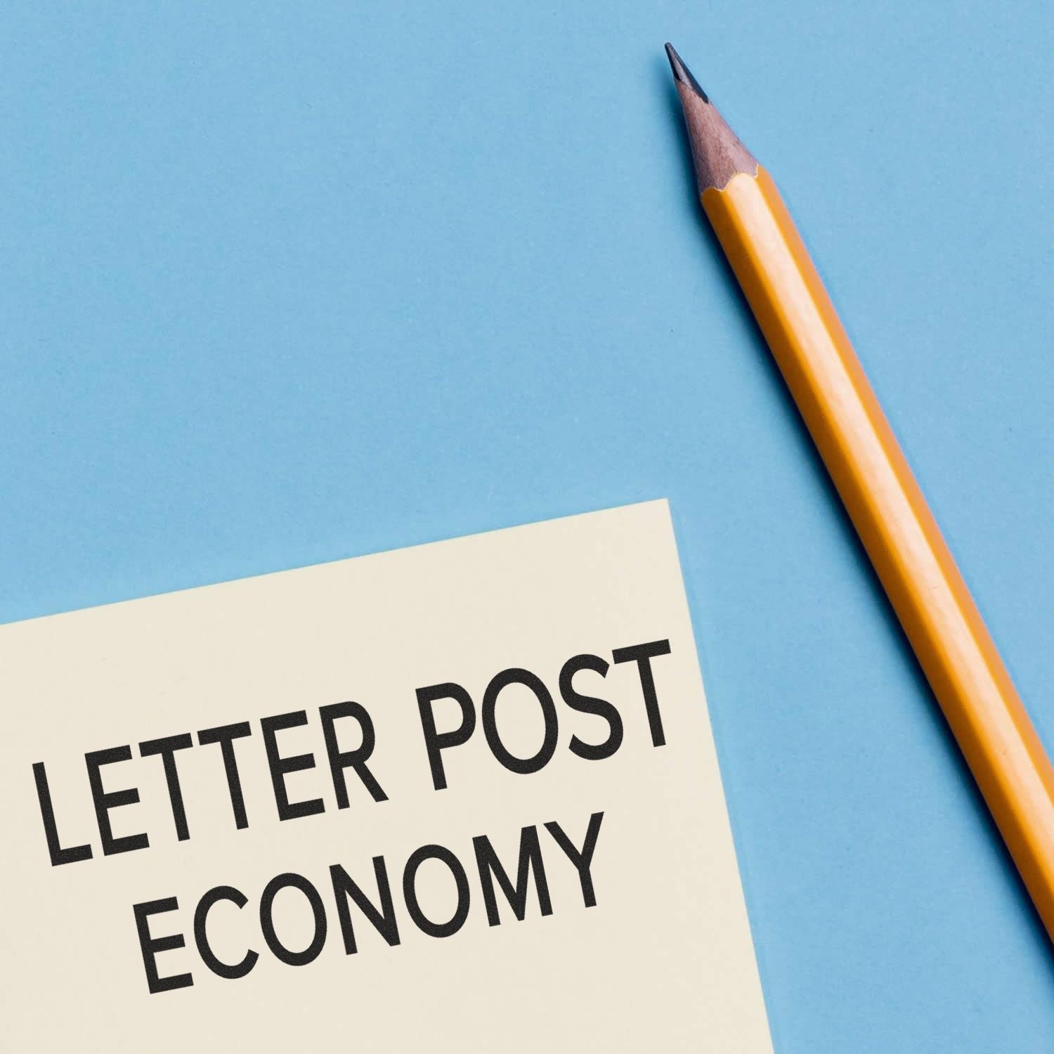 Letter Post Economy rubber stamp impression on paper next to a sharpened yellow pencil on a blue background.