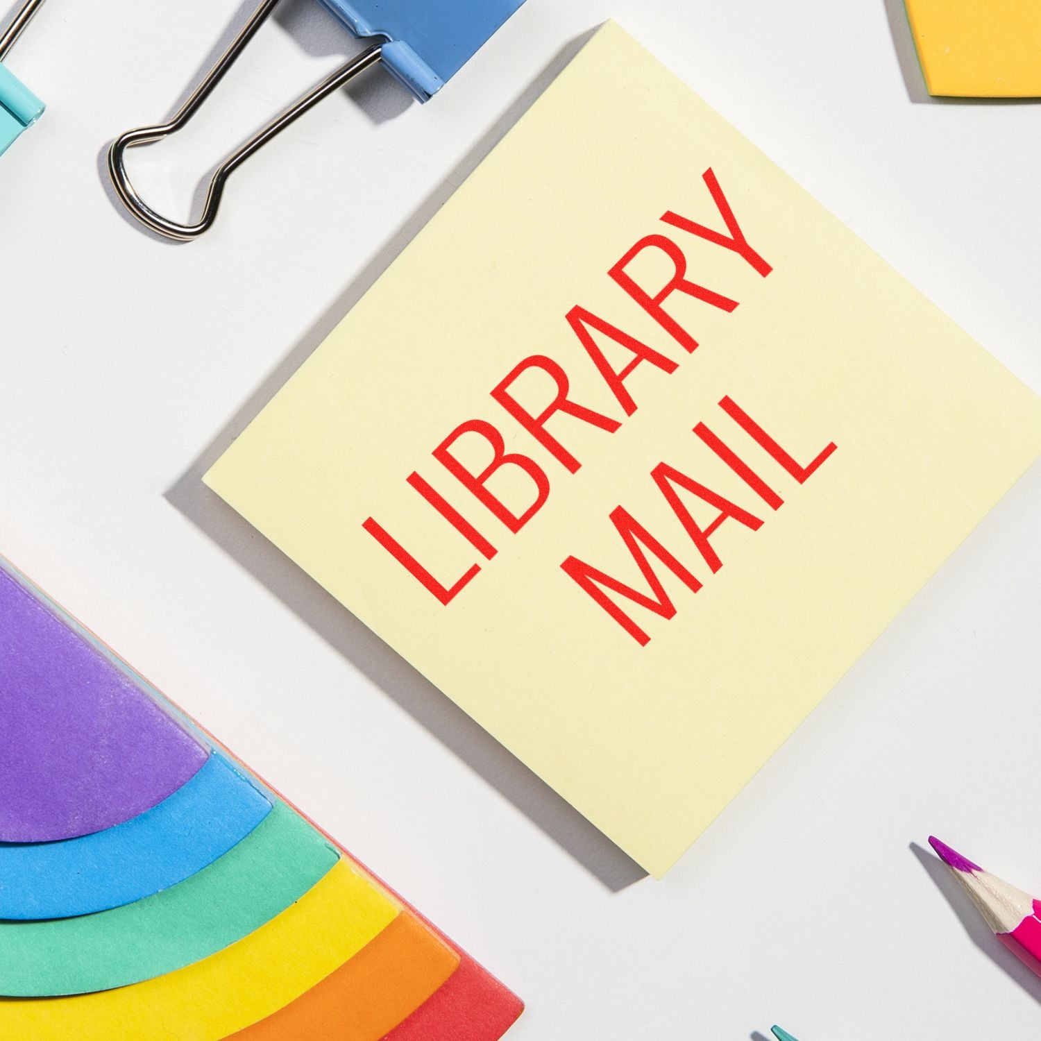 Large Pre-Inked Library Mail Stamp in use on a yellow sticky note, surrounded by colorful office supplies including paper clips and pencils.