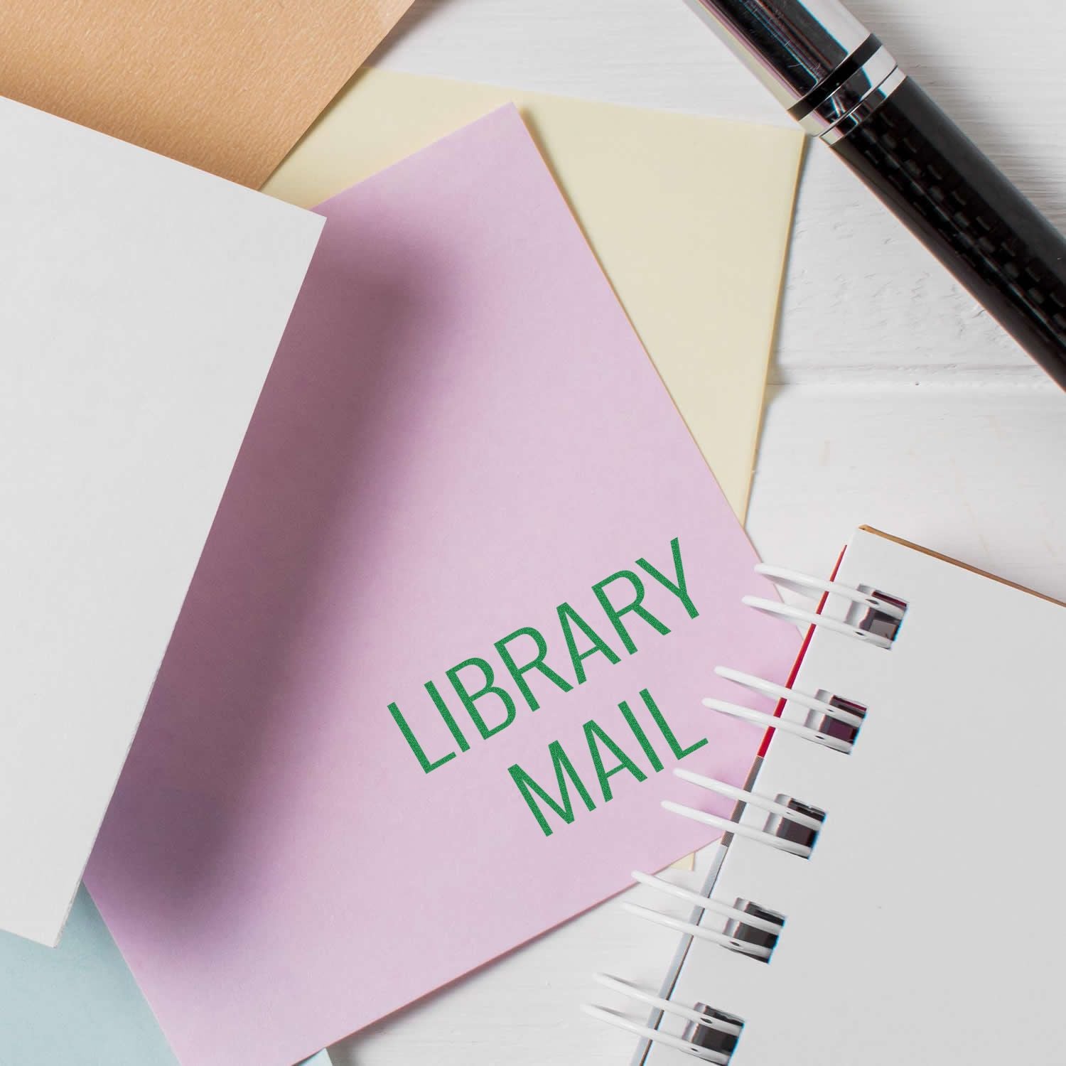 Slim Pre-Inked Library Mail Stamp used on a pink envelope, surrounded by other stationery items and a pen.