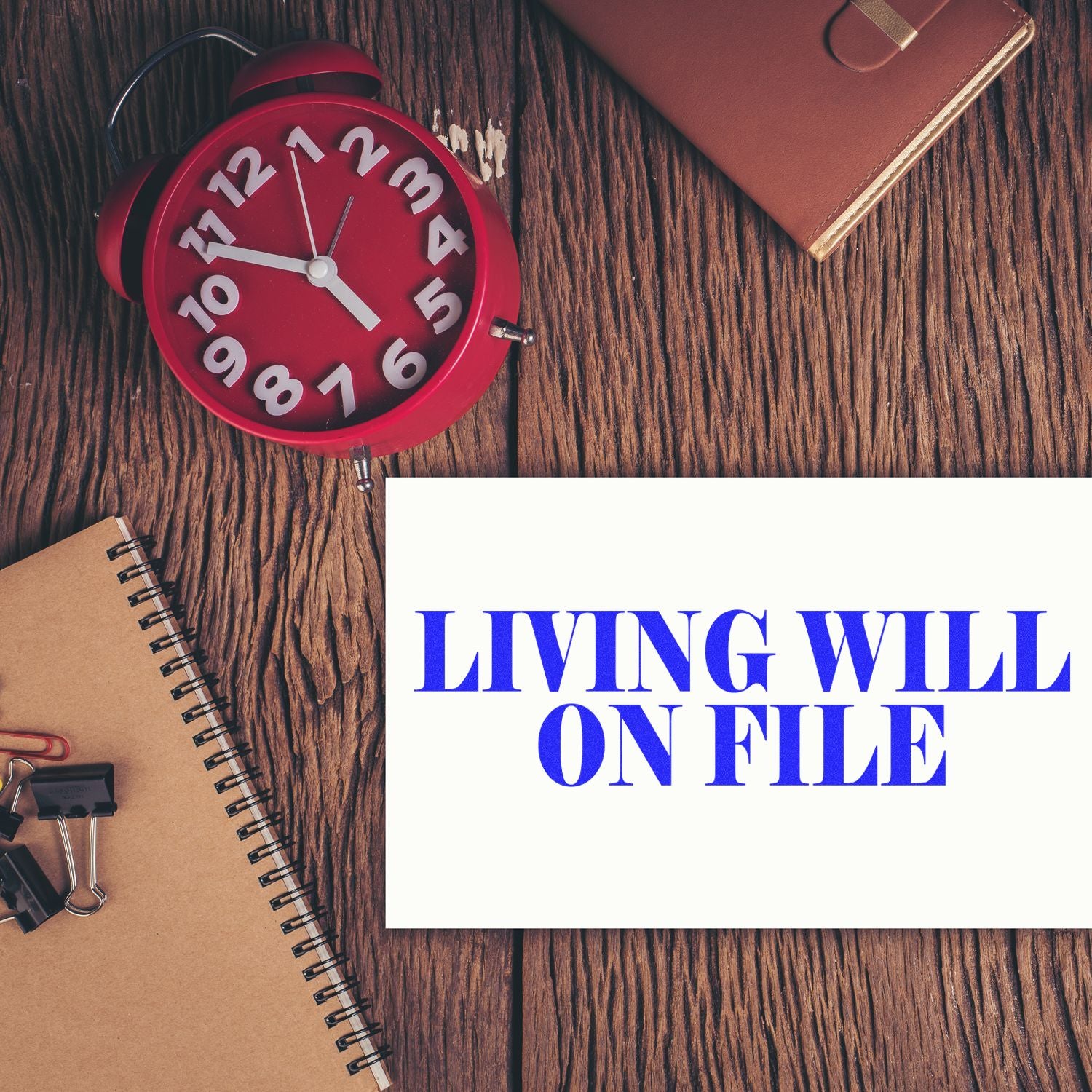 Large Living Will On File Rubber Stamp used on a white paper, placed on a wooden desk with a red clock, notebook, and binder clips.