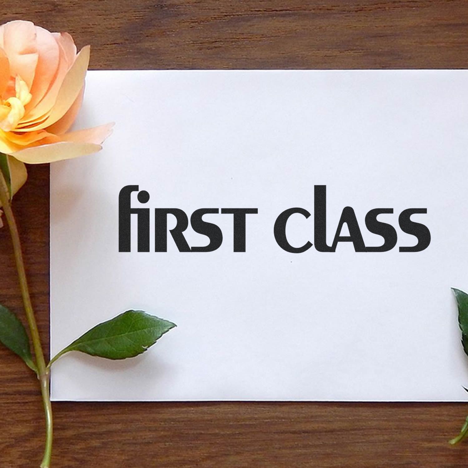 Lower case first class rubber stamp impression on white paper, placed on a wooden surface with a yellow rose and green leaves.