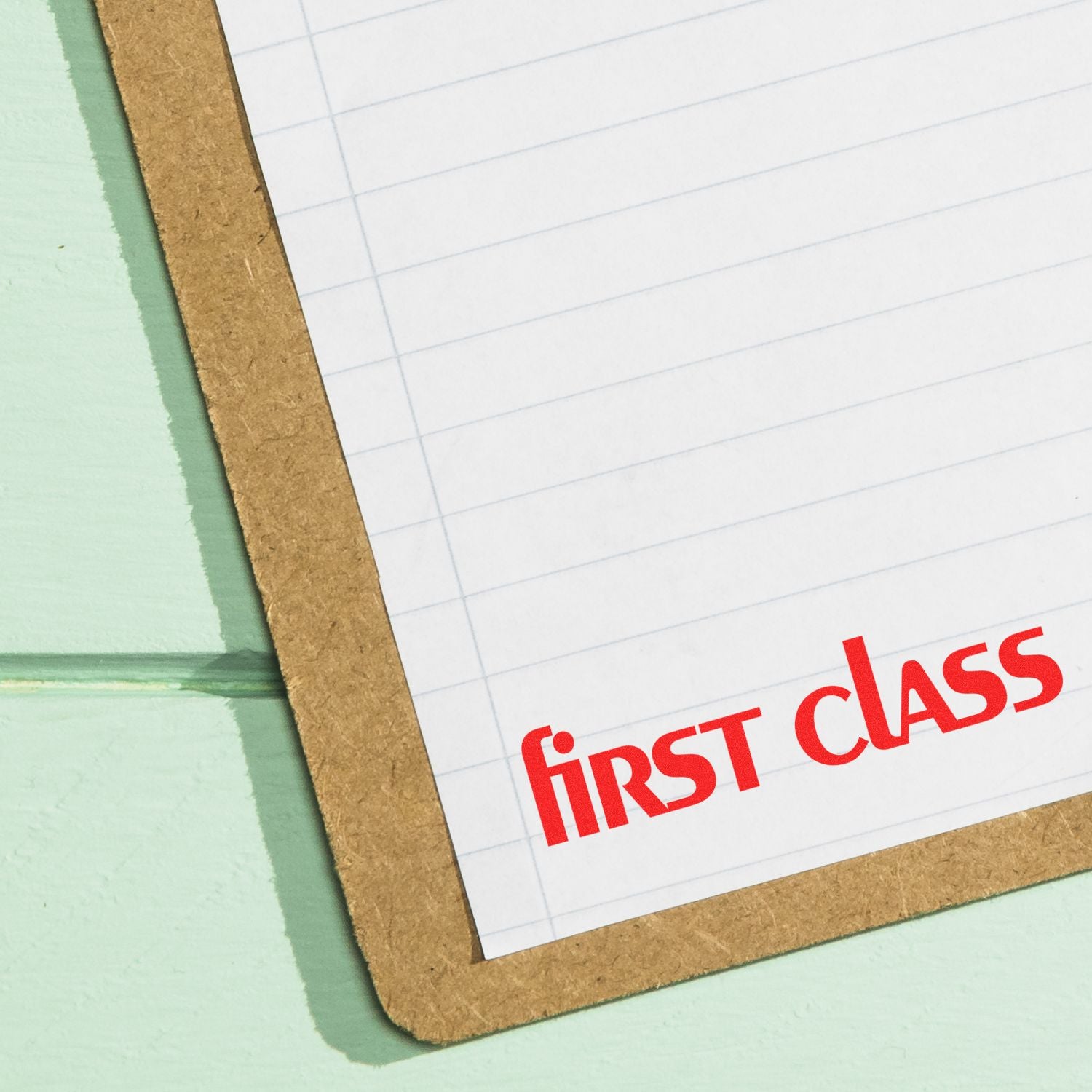 Large Lower Case First Class Rubber Stamp in red ink on a lined paper attached to a brown clipboard, placed on a light green surface.