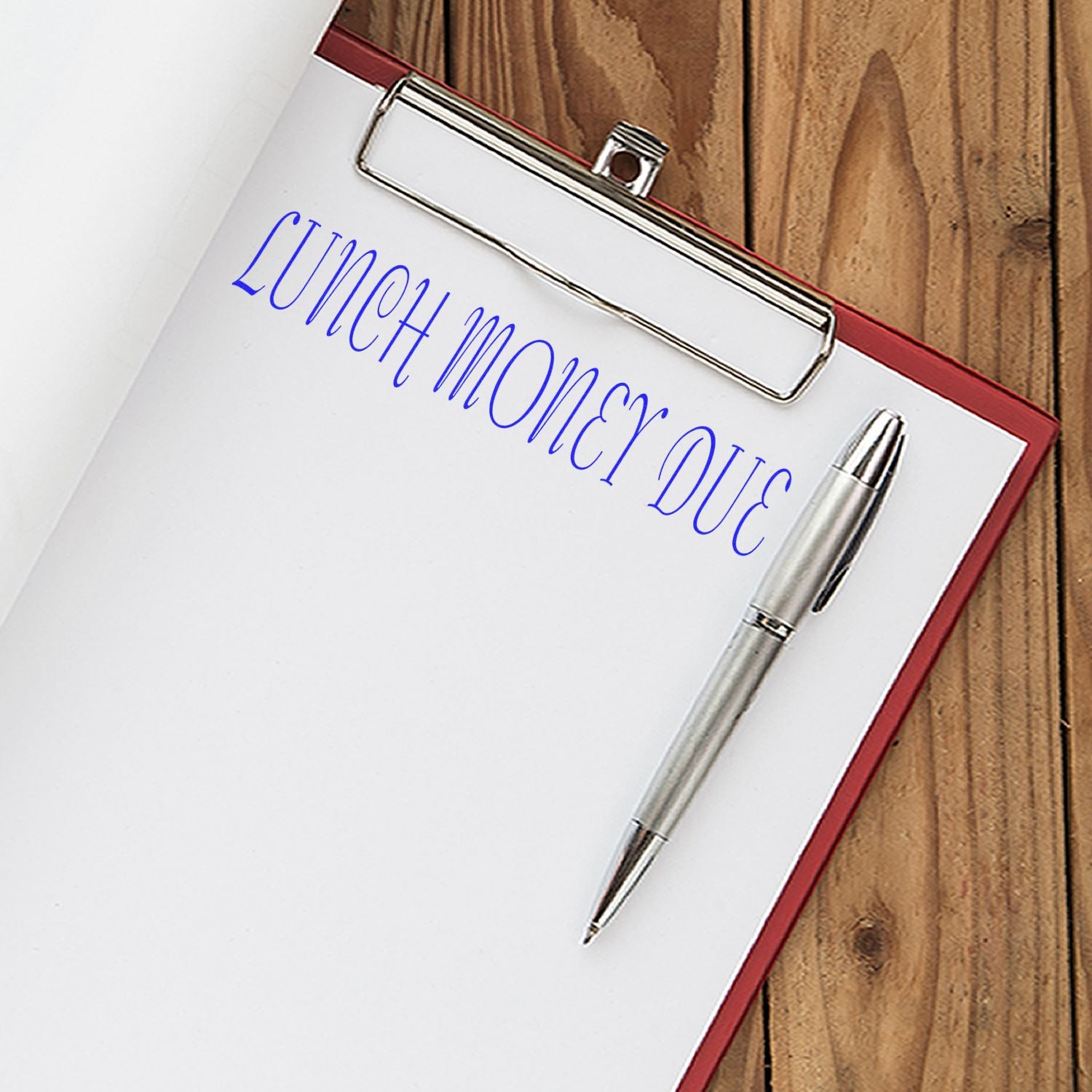 Clipboard with a sheet of paper stamped 'Lunch Money Due' in blue ink, accompanied by a silver pen on a wooden surface.