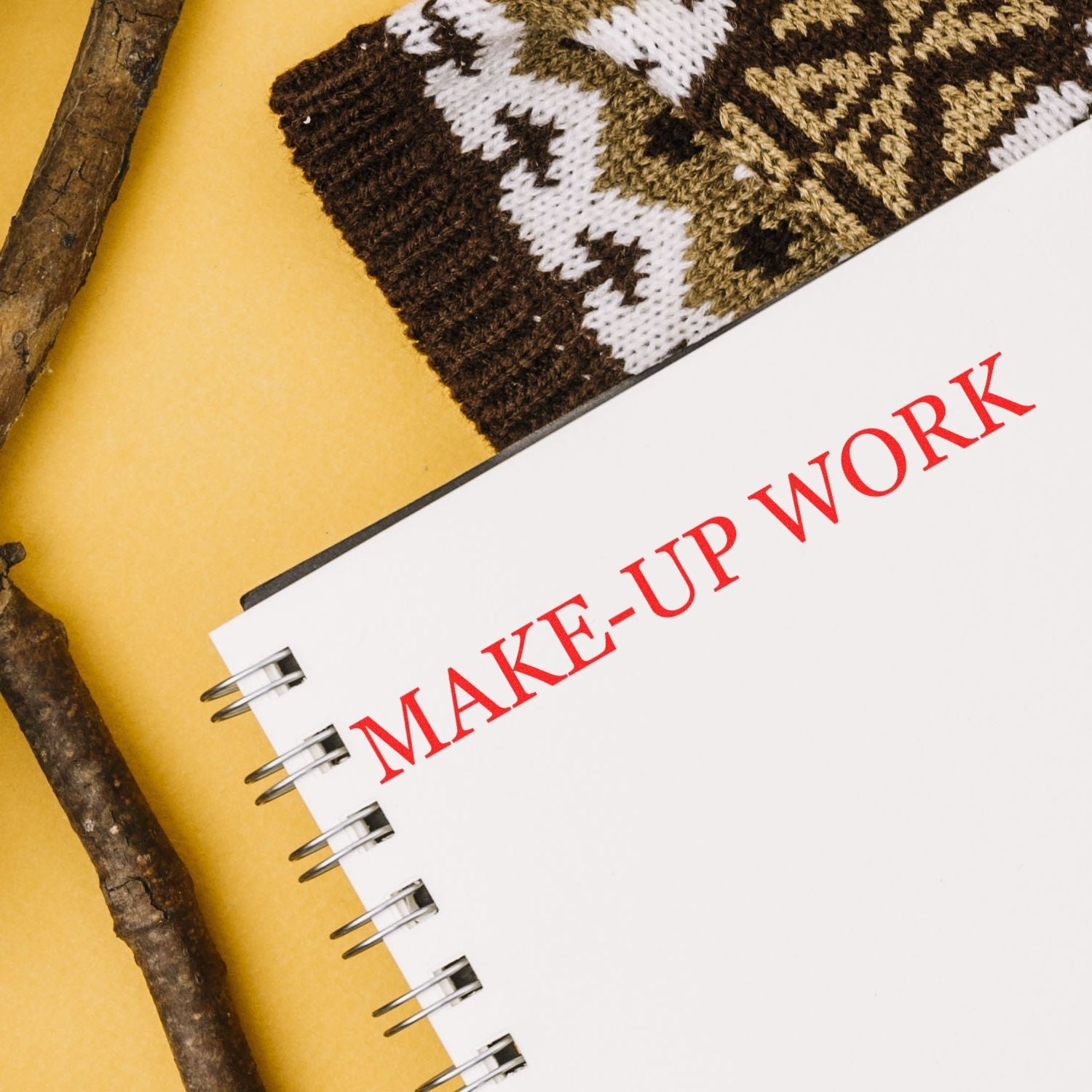 Self Inking Make Up Work Stamp in red ink on a spiral notebook, placed on a yellow surface with a knitted item and wooden sticks nearby.