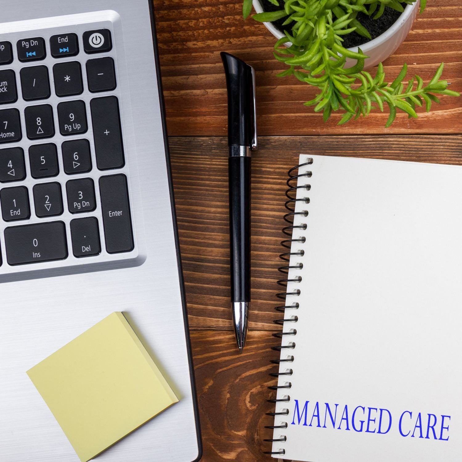 Desk with a laptop, pen, sticky notes, plant, and a notebook stamped with 'Managed Care' in blue ink.