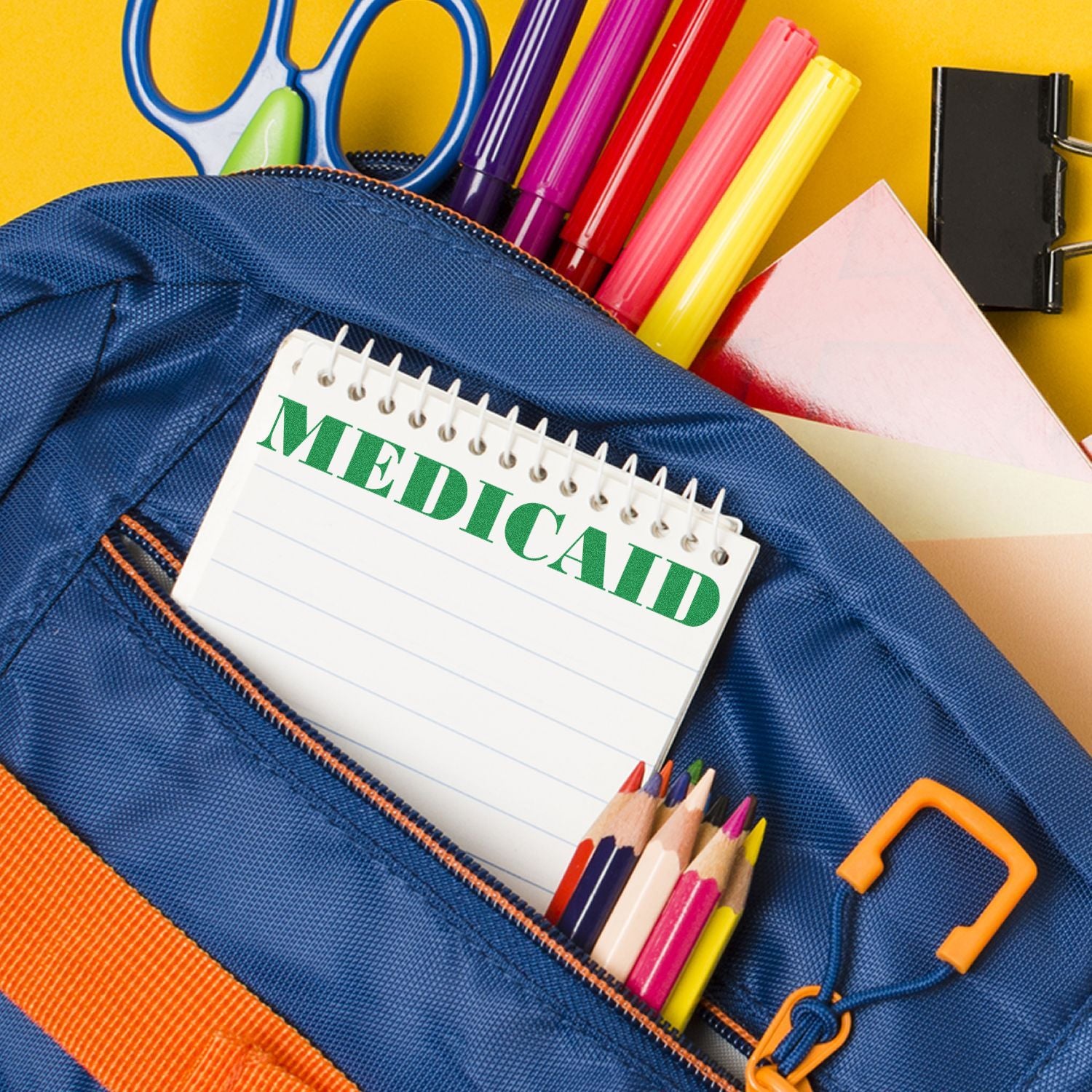 A Medicaid rubber stamp on a notepad inside a blue backpack with colorful pens, pencils, and scissors on a yellow background.