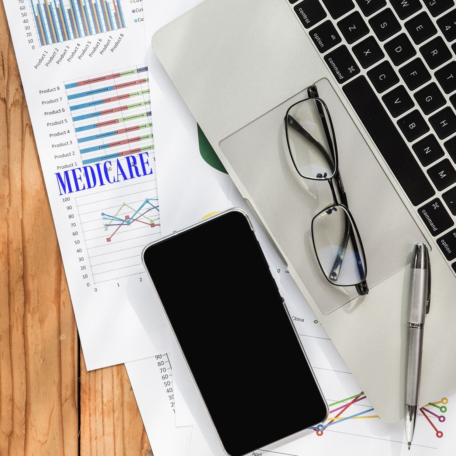 Self Inking Medicare Stamp on a document with charts, beside a laptop, glasses, smartphone, and pen on a wooden desk.