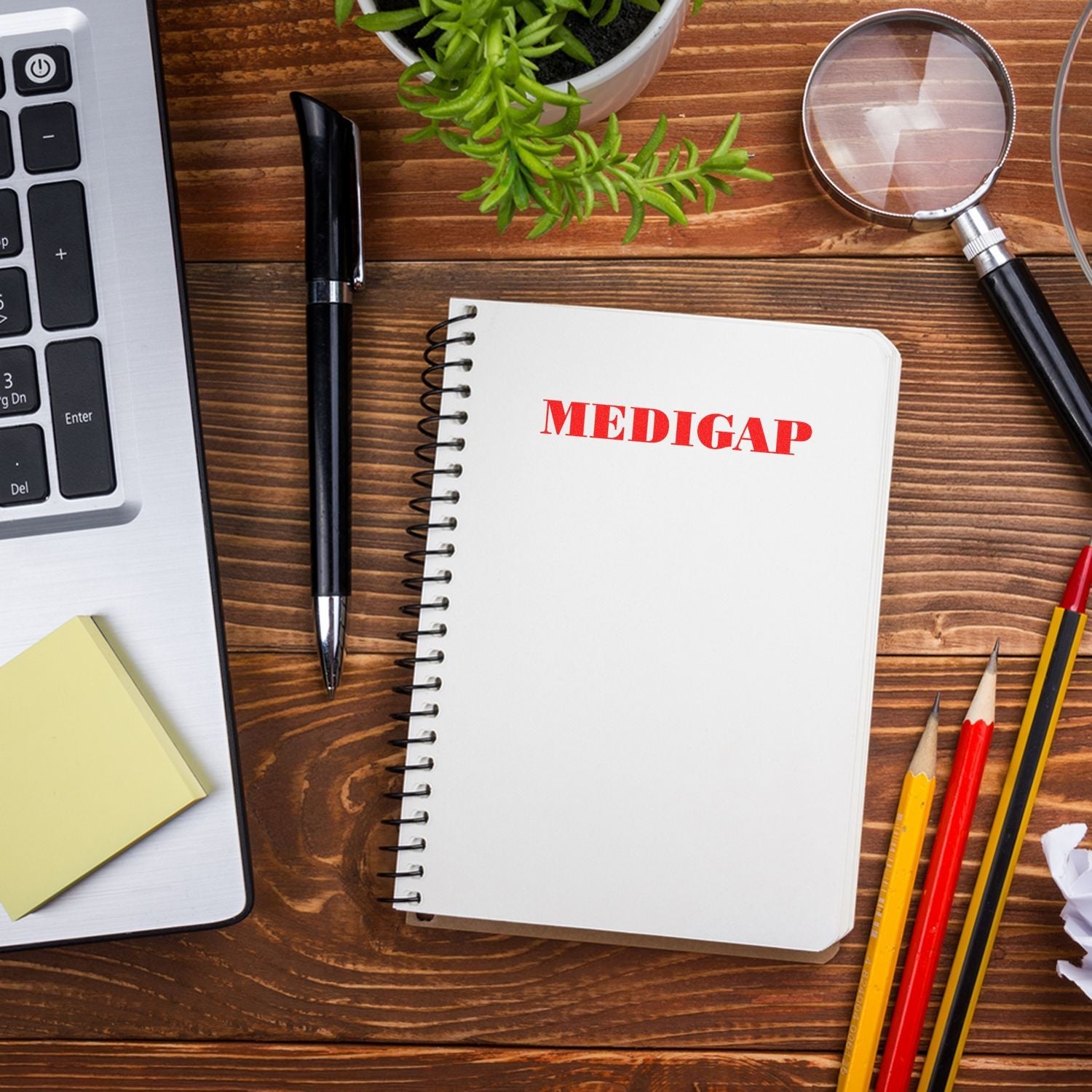 Self Inking Medigap Stamp in use on a spiral notebook, surrounded by office supplies including a laptop, pen, magnifying glass, and plant.