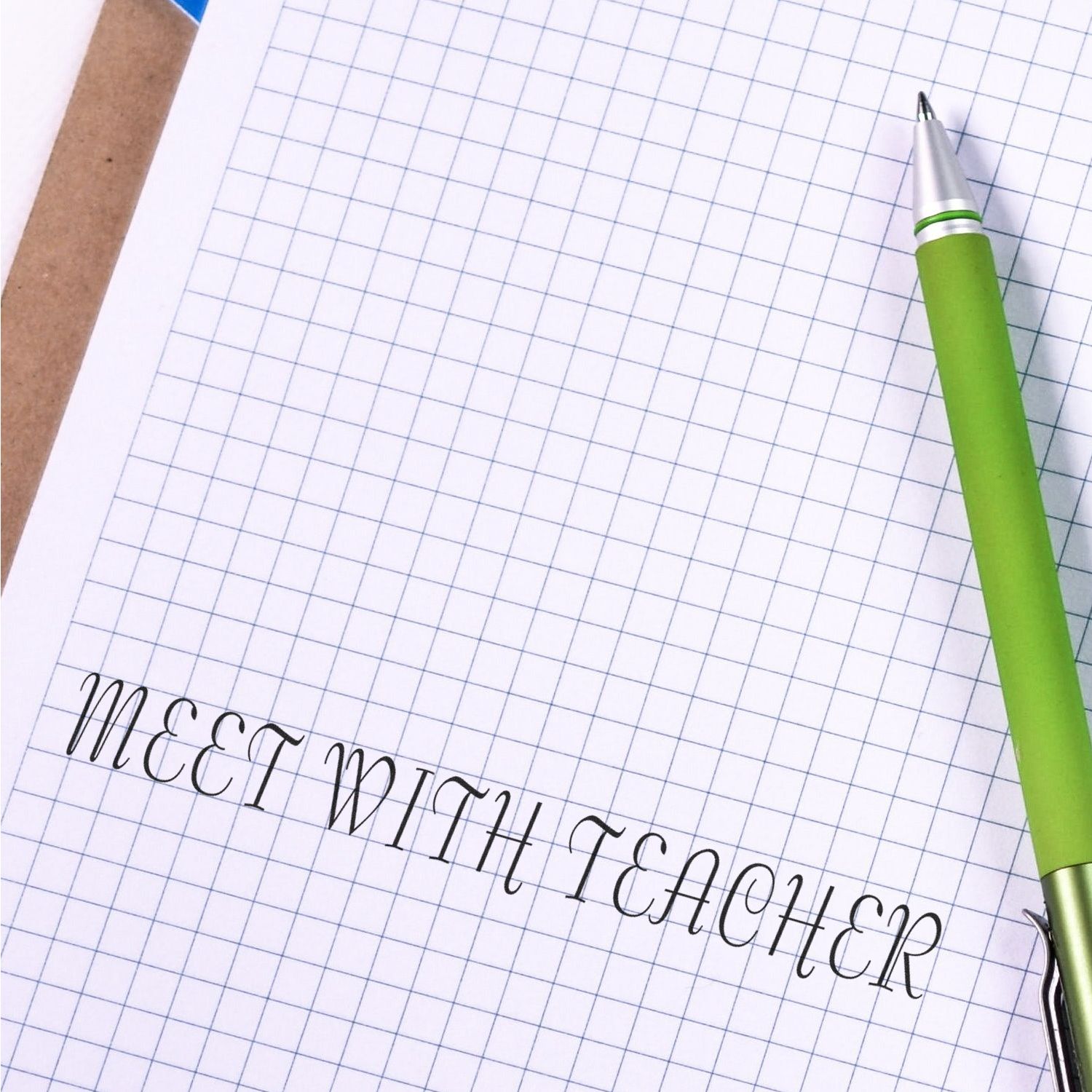 Grid notebook with MEET WITH TEACHER stamped using the Self Inking Meet With Teacher Stamp, next to a green pen.