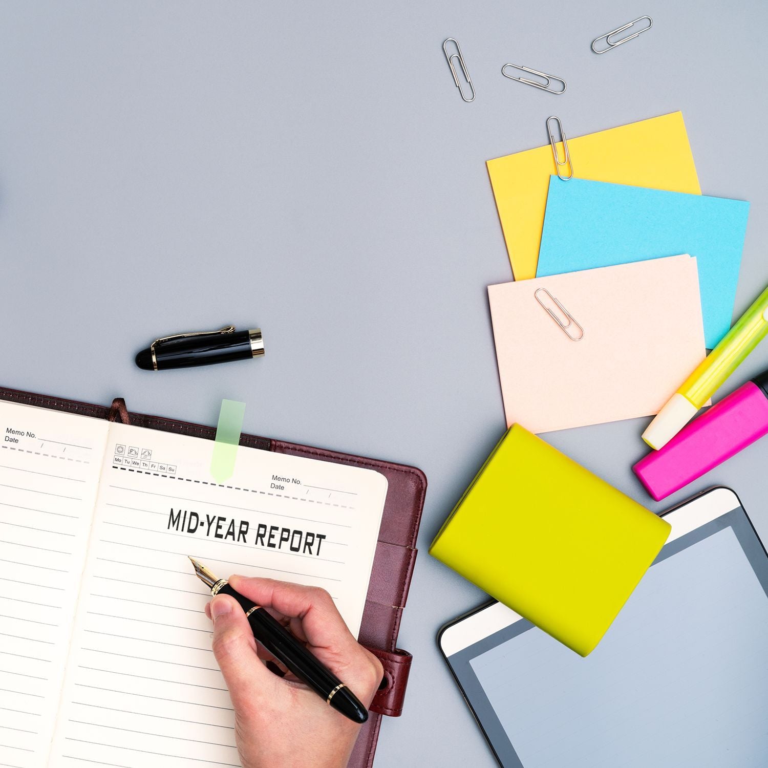 Hand writing in a notebook with a Mid-Year Report rubber stamp, surrounded by colorful stationery and a tablet.