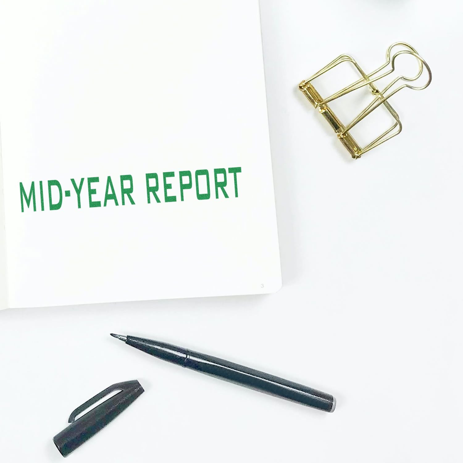 Mid-Year Report rubber stamp on a white paper, with a black pen, pen cap, and a gold binder clip on a white surface.