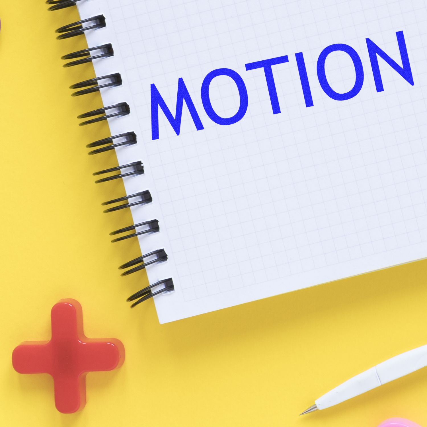 Self Inking Motion Stamp in use on a notebook with the word MOTION stamped in blue ink, surrounded by a red cross and a white pen on a yellow background.