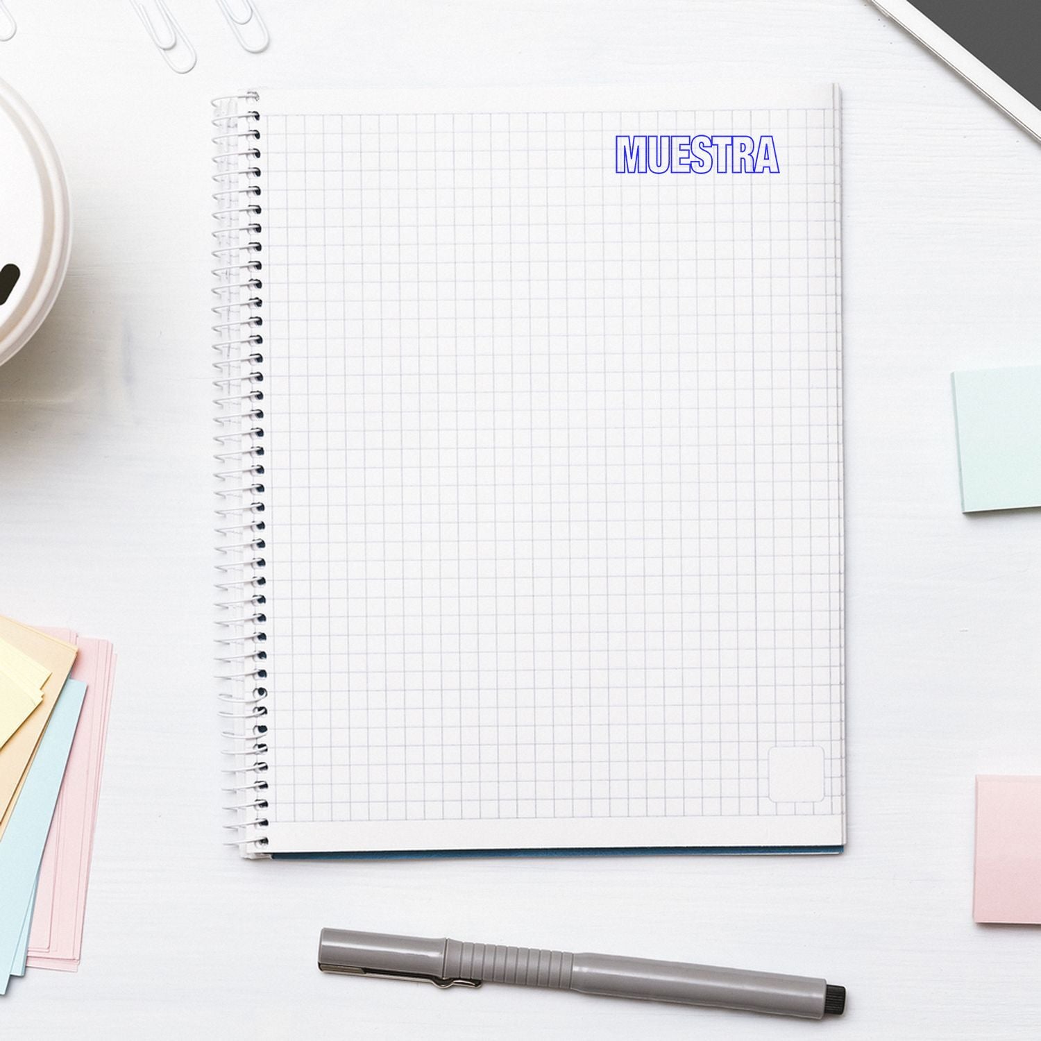 Large Self Inking Muestra Stamp used on a spiral notebook with grid paper, surrounded by office supplies on a white desk.