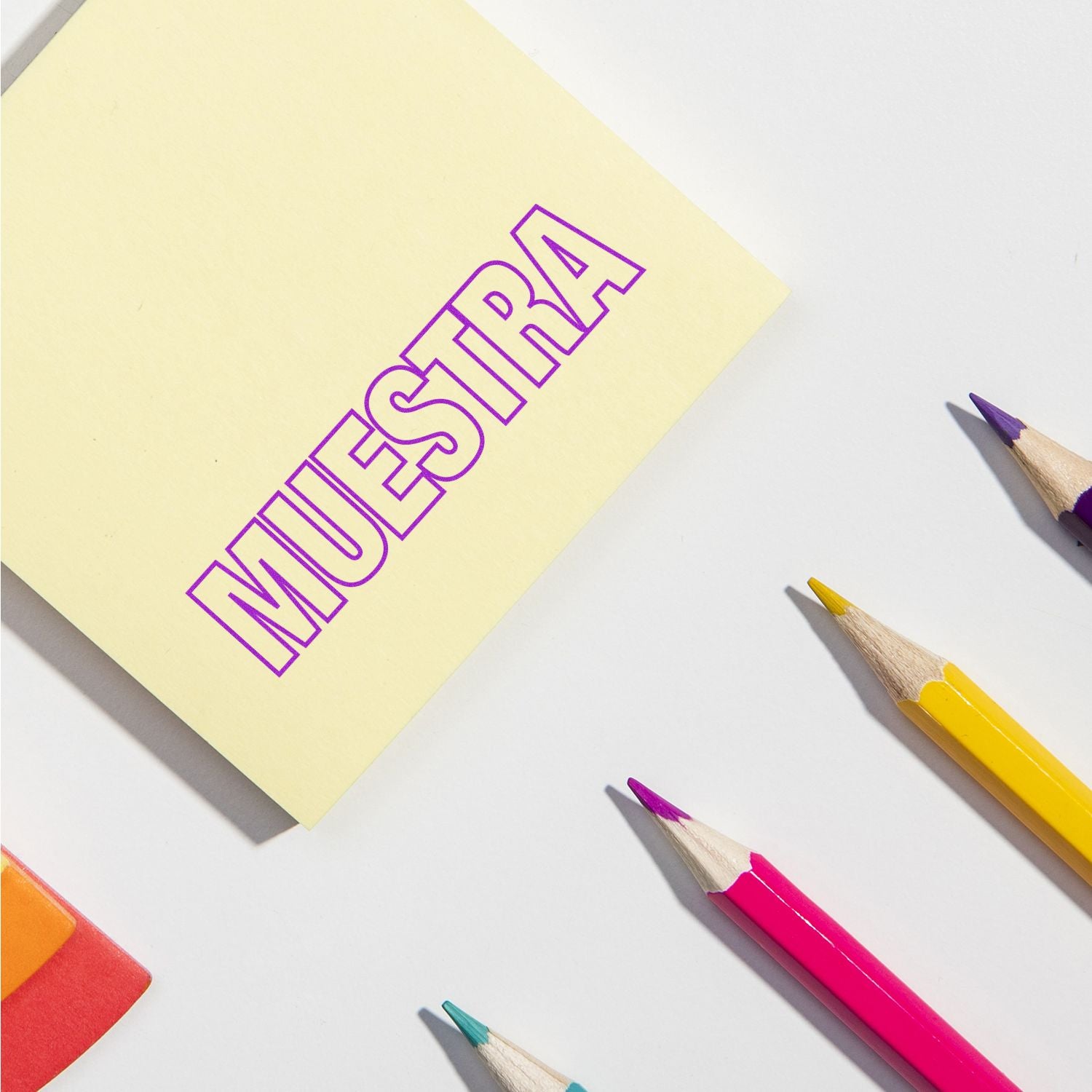 Sticky note with MUESTRA stamped in pink ink using a Muestra Rubber Stamp, surrounded by colorful pencils and an eraser.