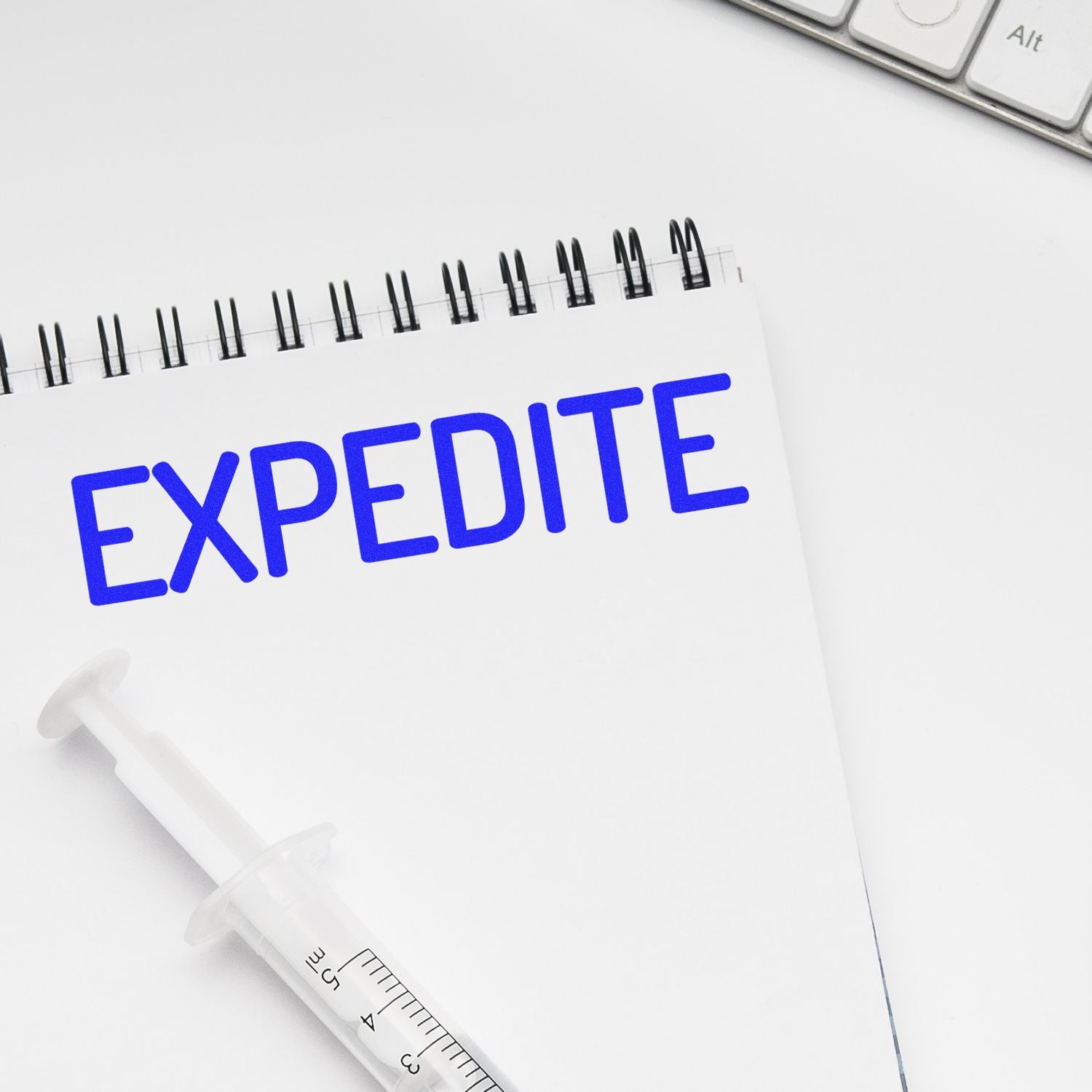 Narrow Expedite rubber stamp marking the word EXPEDITE in blue ink on a white notepad, with a syringe pen nearby.