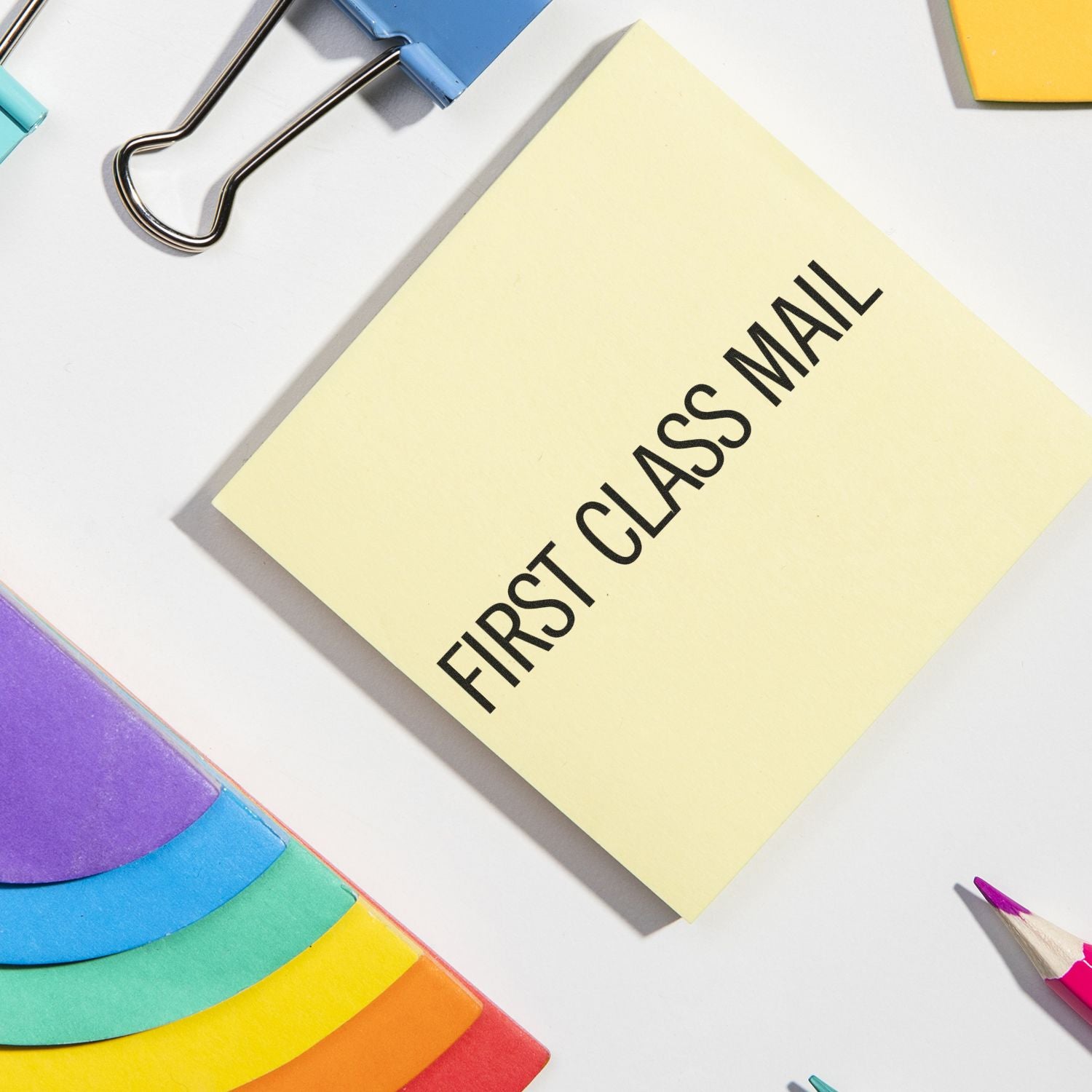 Narrow First Class Mail rubber stamp on a yellow sticky note, surrounded by colorful folders, a binder clip, and pencils.