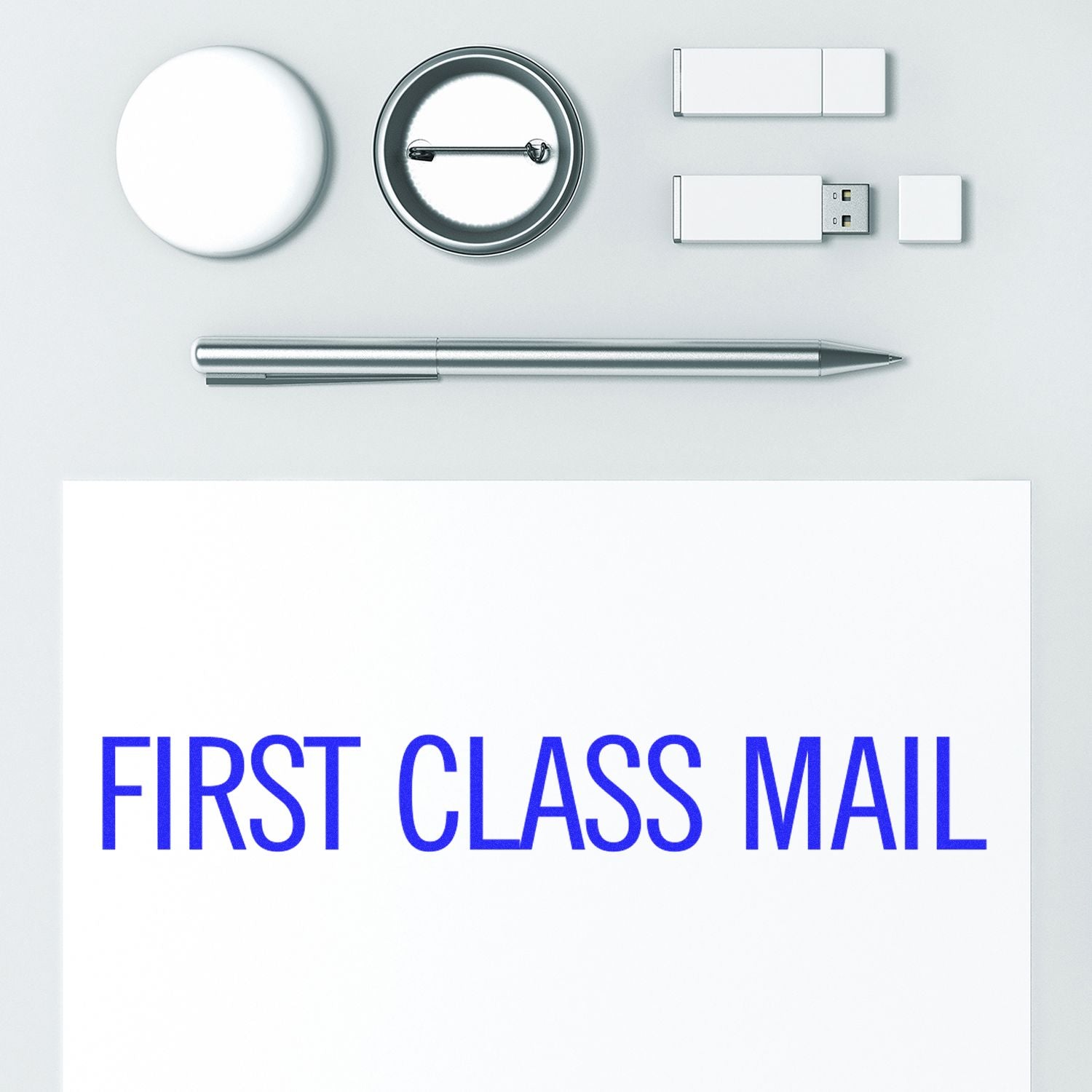 Narrow First Class Mail rubber stamp on white paper, surrounded by a pen, USB drives, a button, and a round object.