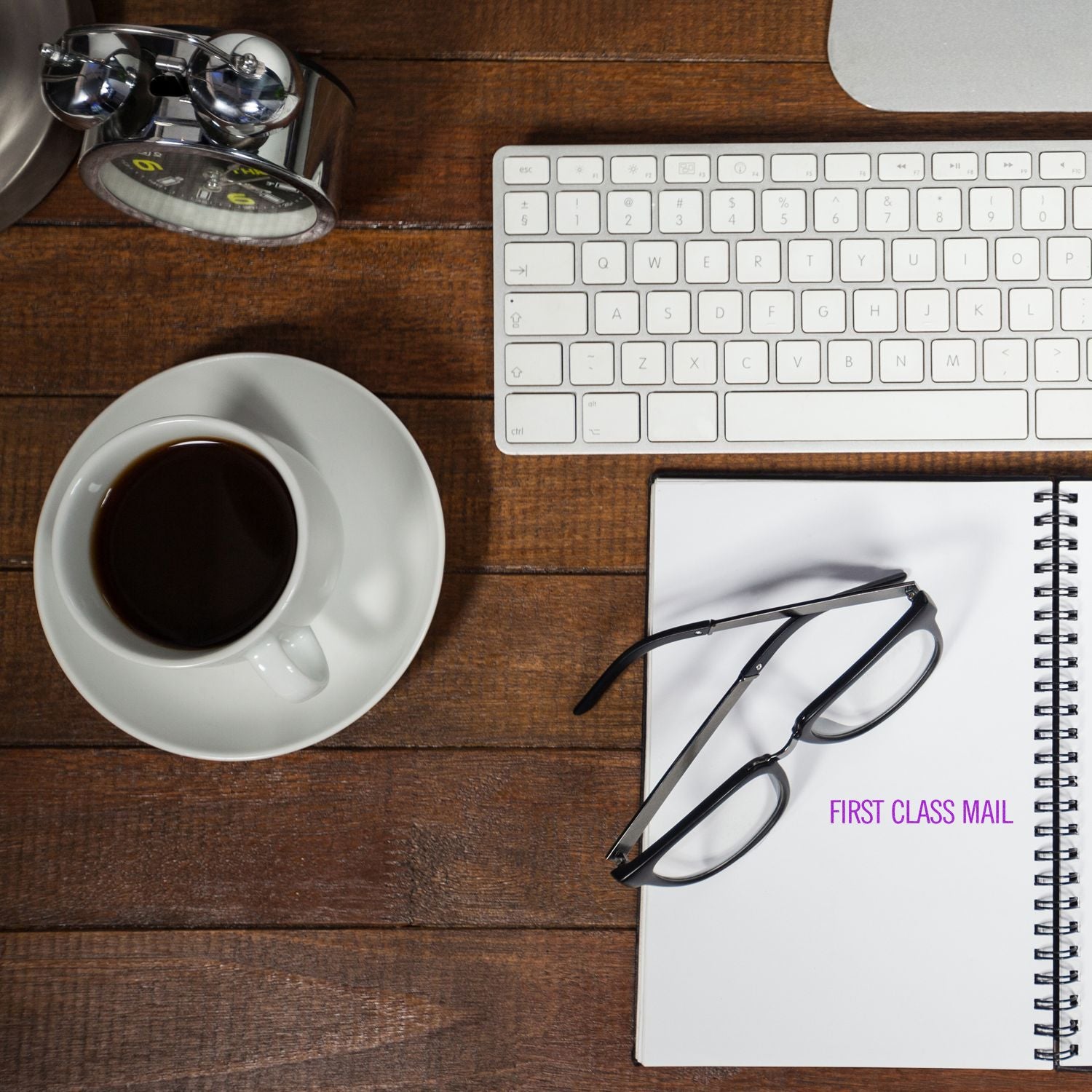 Narrow First Class Mail rubber stamp on a notebook beside glasses, coffee cup, keyboard, and clock on a wooden desk.